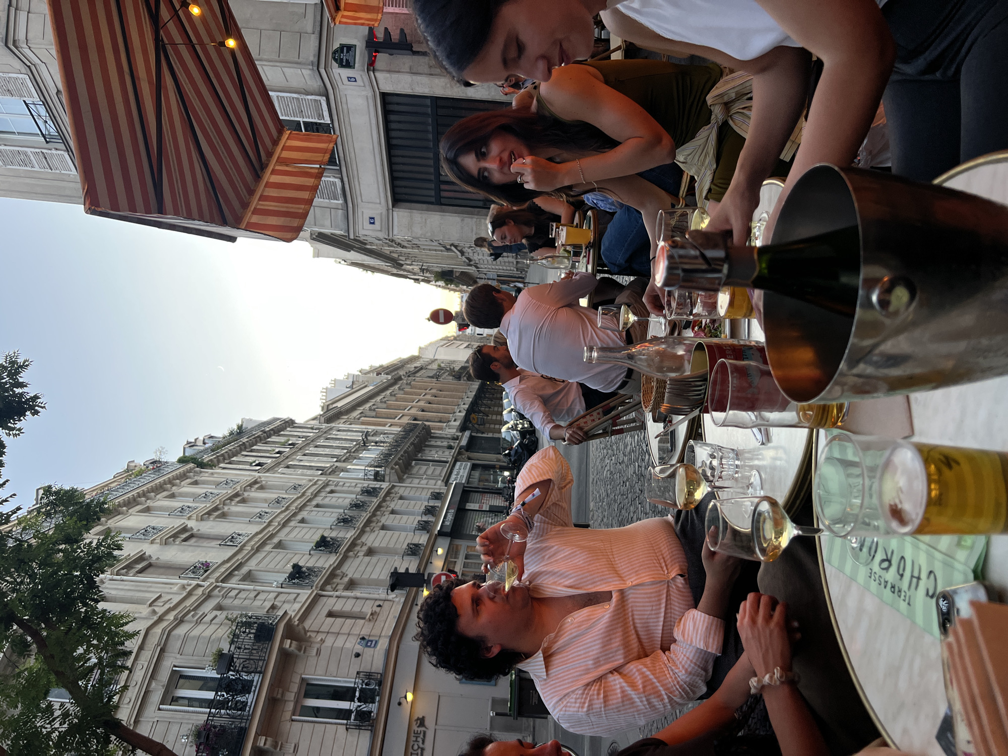 In Paris, France, on a cobblestone street lined with classic Haussmannian architecture, a group of people are dining at an outdoor cafe. The late afternoon sun casts a warm glow on the scene. In the foreground, a man with dark, curly hair is drinking from a wine glass. He is wearing a light pink striped shirt. To his right, a woman with dark hair is looking to her left and touching her chin. In front of them on the table are glasses of wine and beer, a bottle of champagne in an ice bucket, and menus. In the background, other diners are seated at tables, some conversing, others looking out at the street. The cafe's red and white striped awning provides shade. The atmosphere is relaxed and social, indicative of a typical Parisian cafe experience.