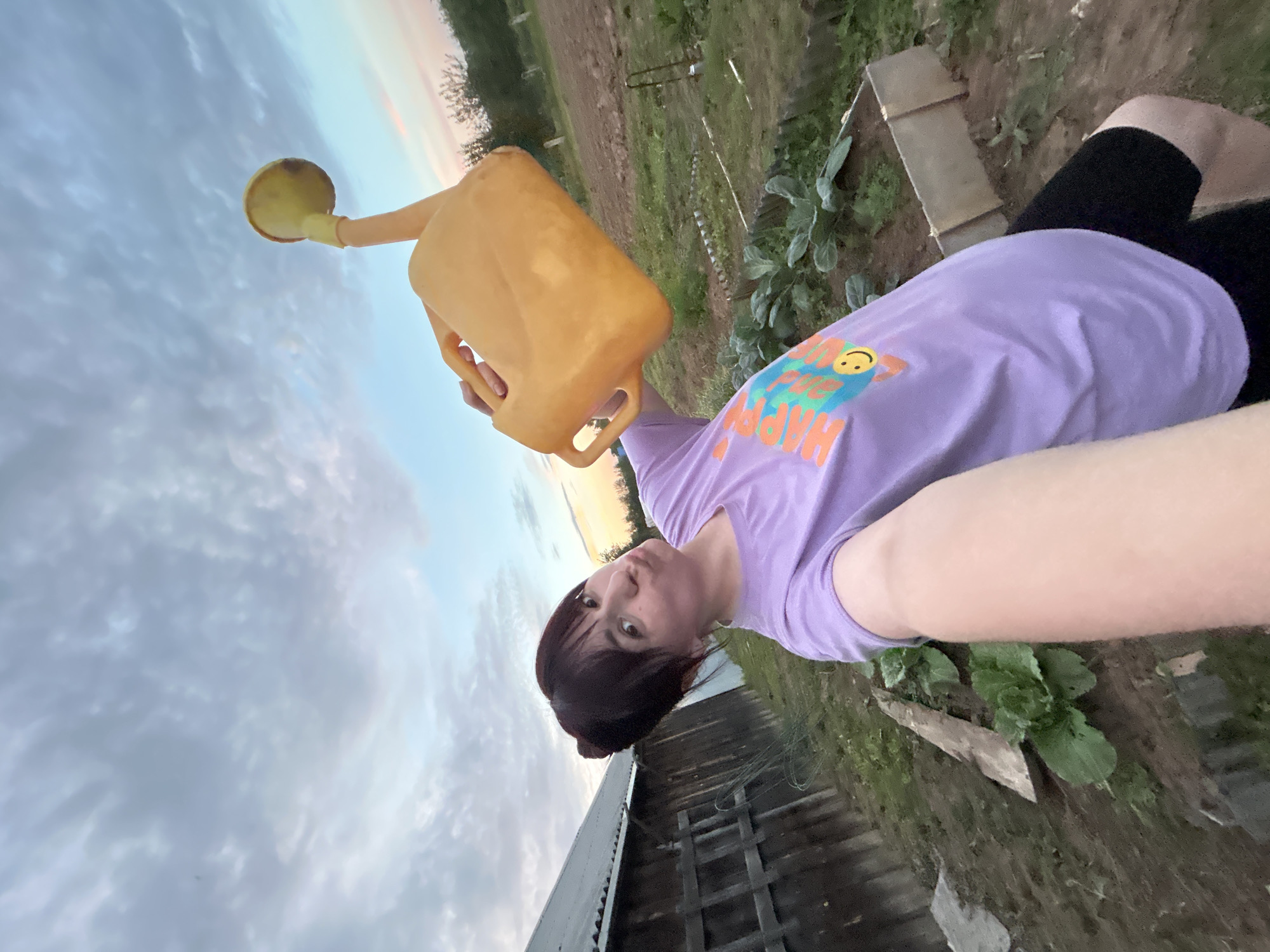 The image is a first-person perspective selfie taken outdoors in what appears to be a rural or garden setting. A woman with dark hair pulled up is in the foreground, holding a yellow watering can as if to pour it. She is wearing a purple t-shirt with a colorful graphic of a smiley face. Behind her, to the left, is a wooden structure resembling a shed or outbuilding with a corrugated metal roof. The ground is dirt and grass, with some garden beds containing leafy green plants. In the background, there is more open ground, some trees, and a cloudy sky with hints of sunset colors. The location is identified as Фоки, Russia.