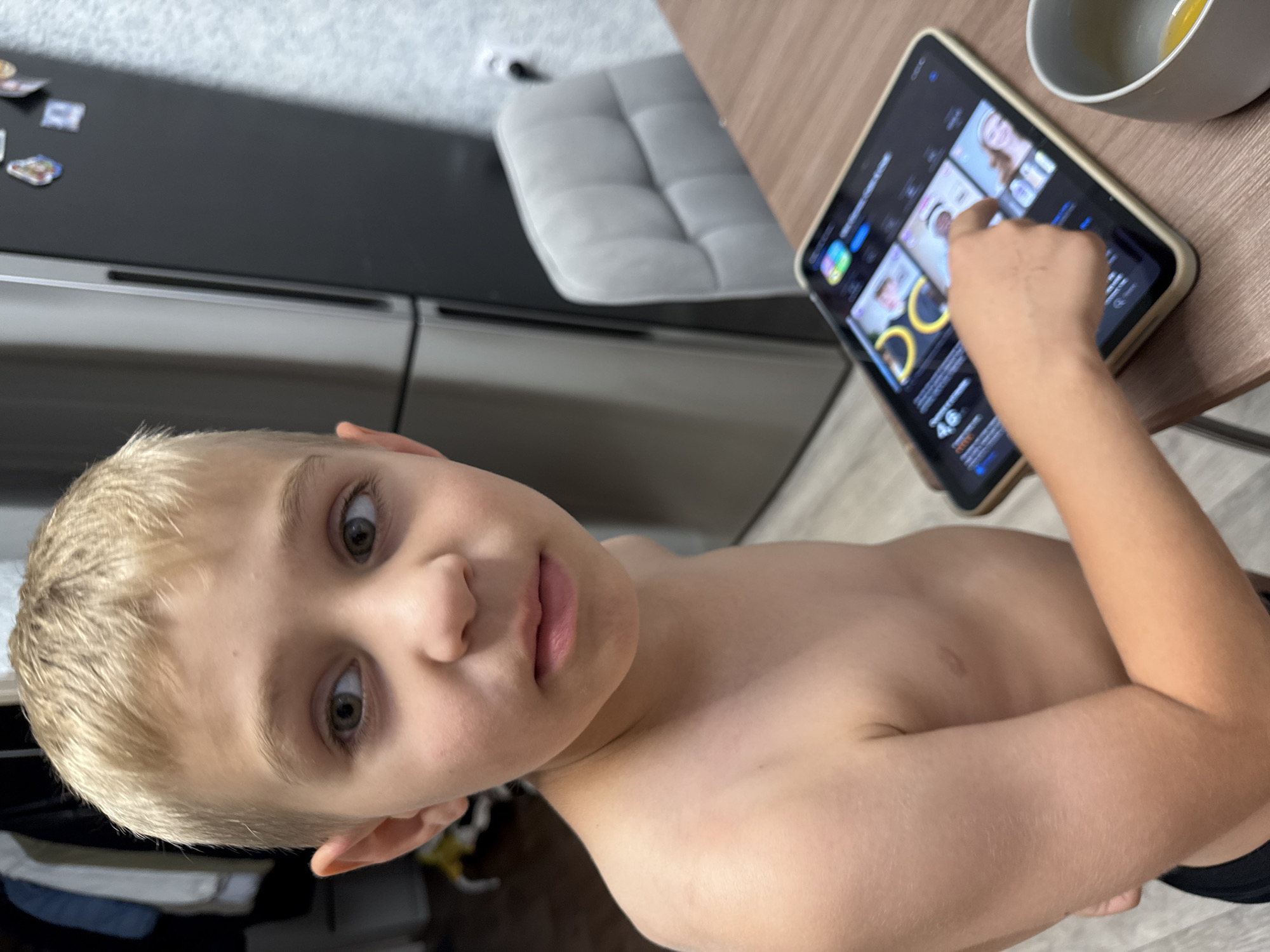 A close-up shot captures a young boy with blonde hair and light green eyes, looking directly at the camera with a curious expression. He is shirtless, revealing his chest and shoulder. His right arm is extended, with his finger touching a tablet that rests on a wooden table. The tablet displays a screen with various images and text, appearing to be an application or website. To the right of the tablet, a white bowl with a yellow liquid, possibly food or drink, is partially visible. In the background, a gray tufted chair is visible, and a kitchen counter with magnets is on the left. The overall scene suggests a casual indoor setting, possibly a kitchen or dining area. The lighting appears to be natural, indicating daytime.