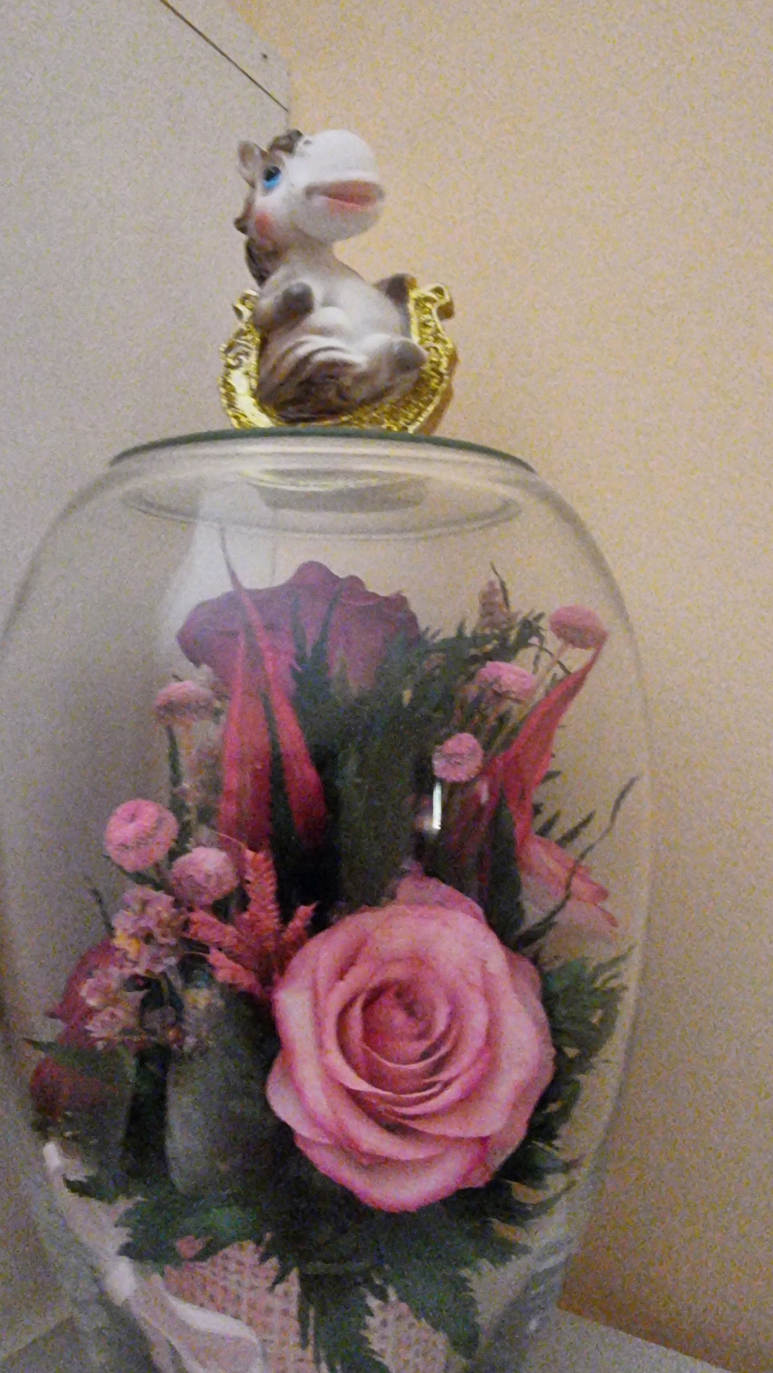 An ornamental glass dome contains a pink rose bouquet and a small figurine of a horse. The horse figurine is white with blue eyes and is sitting on a gold-colored decorative base. The bouquet consists of a large pink rose, smaller pink flowers, and dark green foliage. The glass dome is placed on a surface in front of a beige wall. The image appears to be a close-up shot of a decorative display.
