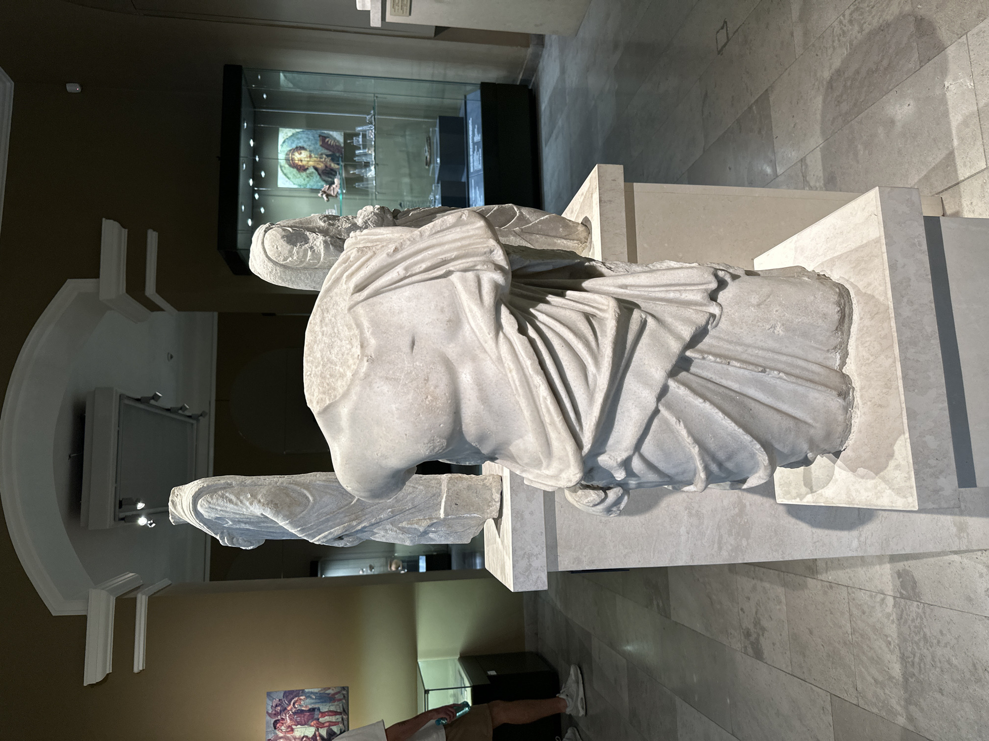 The image depicts a museum exhibit in Varna, Bulgaria, featuring ancient marble sculptures. In the foreground, a headless torso of a draped statue is prominently displayed on a marble pedestal. To its left and slightly behind, another full-body draped statue stands on a similar pedestal. Further back, to the right, a glass display case showcases various artifacts, with a circular framed artwork featuring a person in historical attire visible within. The museum environment includes a tiled floor and muted brown walls, with architectural elements like arches and recessed lighting visible in the ceiling. A person, partially visible on the left, is wearing a white t-shirt and khaki shorts, holding a blue bottle. The scene captures a moment within a museum setting, highlighting the ancient art on display.