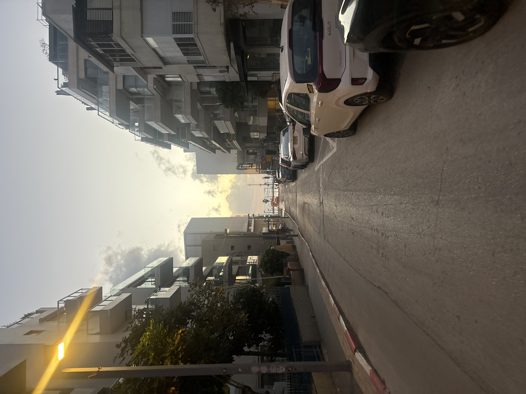 The image depicts a street in Tel Aviv, Israel, with buildings lining both sides. Cars are parked along the right side of the road. The road itself appears to be paved and slightly curved. In the distance, the sea is visible between the buildings. The sky is bright, suggesting it is either early morning or late afternoon. The overall impression is that of a typical residential street in Tel Aviv.