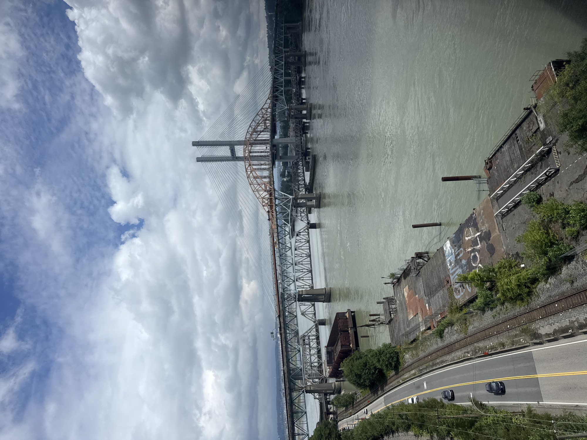The image, taken from an elevated perspective, captures a wide expanse of a river under a cloudy sky. In the foreground, a weathered wooden pier juts out into the murky water, showing signs of disrepair and graffiti. Adjacent to the river is a two-lane road with yellow dividing lines, with several cars driving along it. Next to the road are railway tracks and a dense line of trees. Dominating the midground and background are multiple bridges spanning the river. The most prominent is a large cable-stayed bridge with two tall concrete towers, partially obscured by construction. In front of it, an older steel truss bridge crosses the water, supported by concrete piers. In the distance, the urban landscape of New Westminster, Canada, is visible along the far bank of the river.
