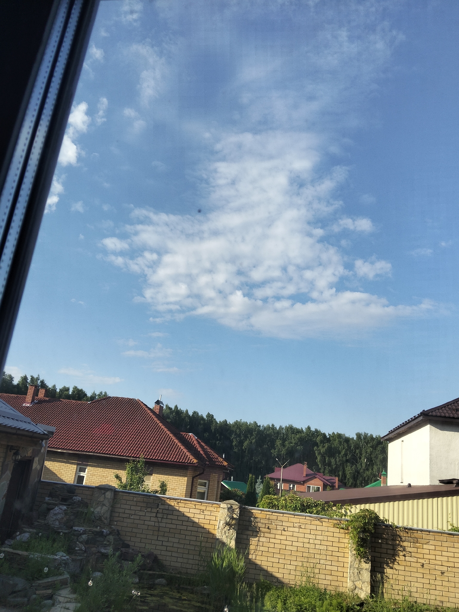 The image shows a view of residential houses and a forest from behind a window. In the foreground, a dark window frame is visible on the left side. The sky is blue with scattered white clouds. Below the sky, there are several houses with red tile roofs and yellow brick walls. A dense line of green trees forms the background. In the lower part of the image, a light brown brick fence and a decorative stone structure with rocks and greenery are visible. The scene appears to be a suburban or rural setting on a clear day.