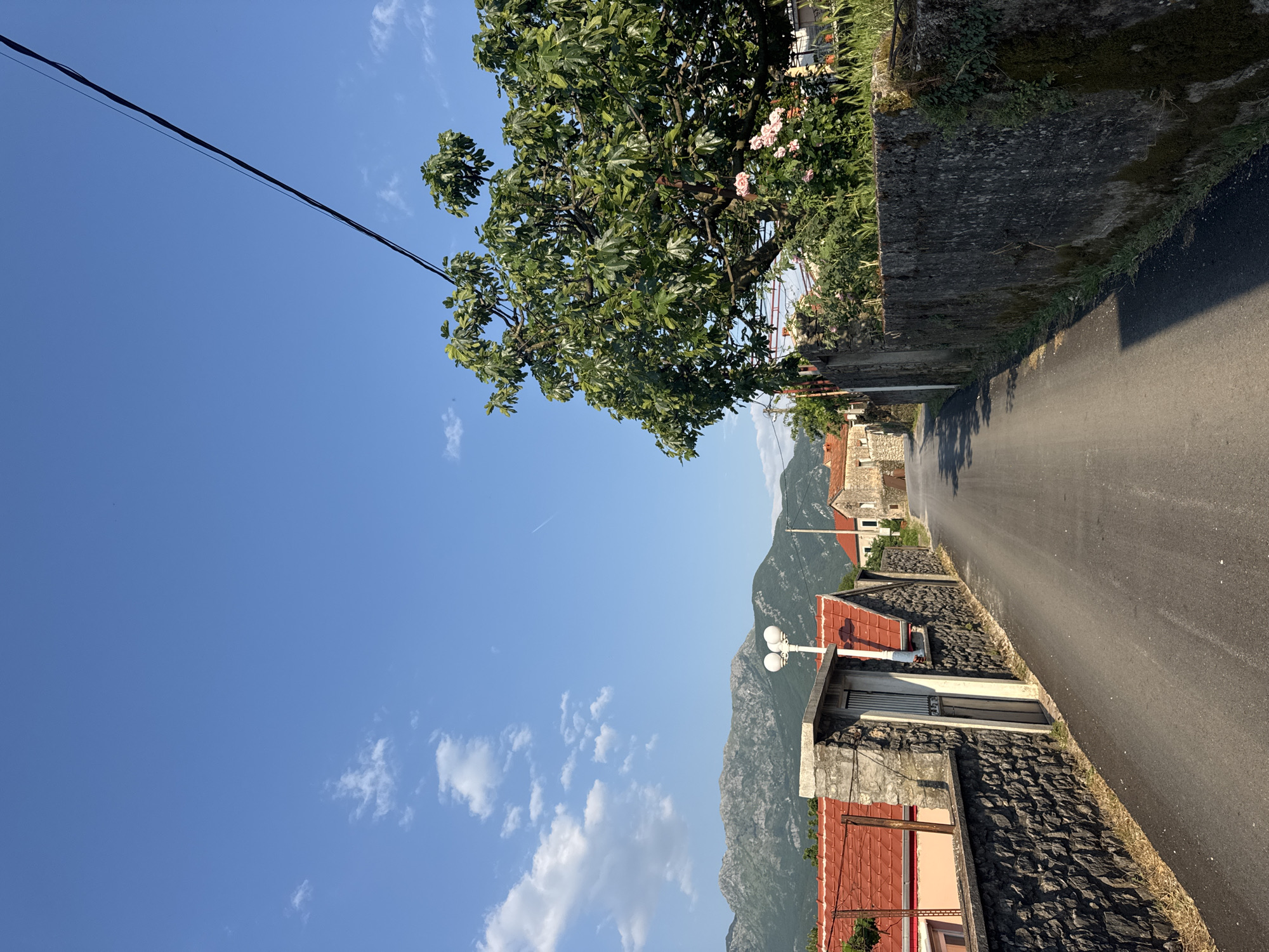 The image shows a narrow asphalt road winding through a residential area in what appears to be the city of Kubasi, Montenegro. The road is flanked by stone walls and buildings with red-tiled roofs. Mountains are visible in the background. The sky is blue with scattered clouds. A tree overhangs the road on the left side. A power line stretches across the upper left of the frame.