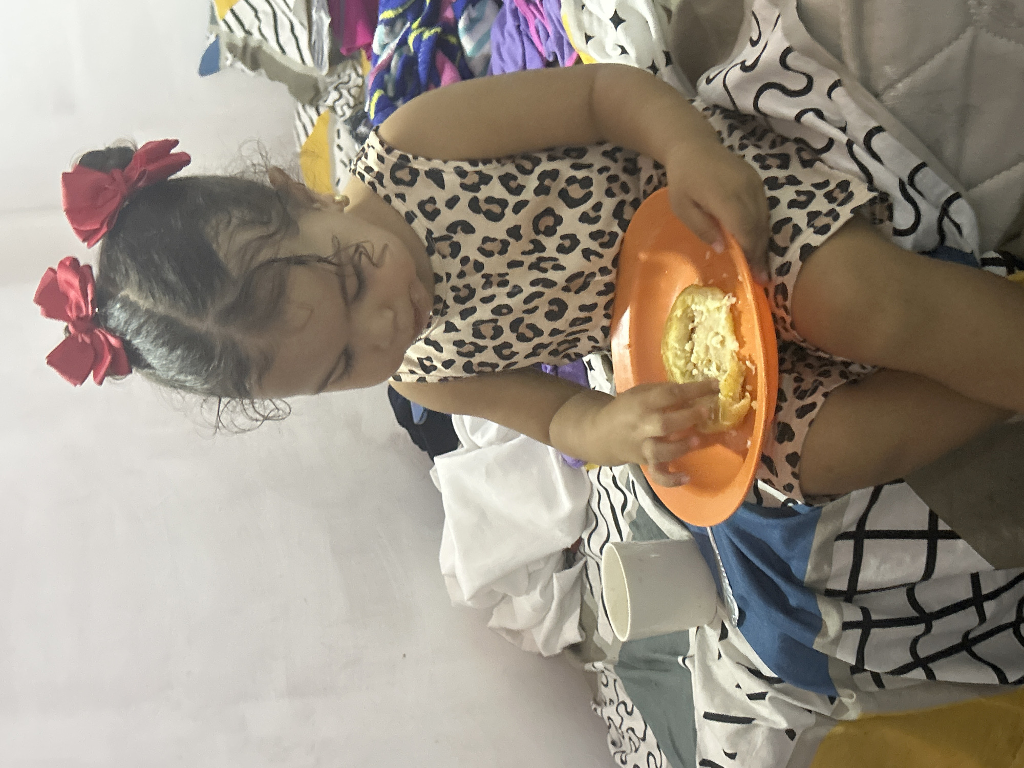 A young child with dark hair pulled into two pigtails adorned with red bows is seated amidst a pile of mixed textiles, including striped and patterned fabrics. The child is wearing a leopard-print garment and holding an orange plate containing a portion of food, which appears to be yellow and creamy. The child is actively eating from the plate with their hands. To the child's left, there is a white cylindrical cup. The setting appears to be indoors with a white wall in the background. There is no discernible information indicating the location is San Juan de Los Morros, Venezuela.
