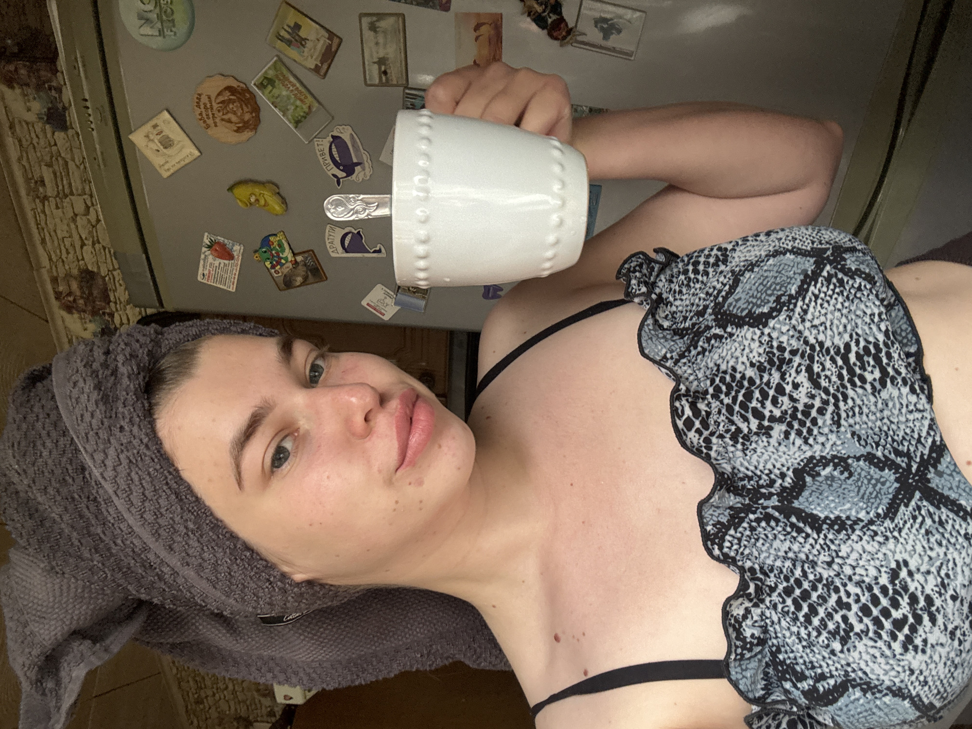 A woman with a towel wrapped around her head holds a white mug with a spoon inside. She is wearing a gray and black snakeskin-print top with black straps. Behind her is a refrigerator covered in magnets. The location in the city of Мещёры, Russia is not discernible from the image.