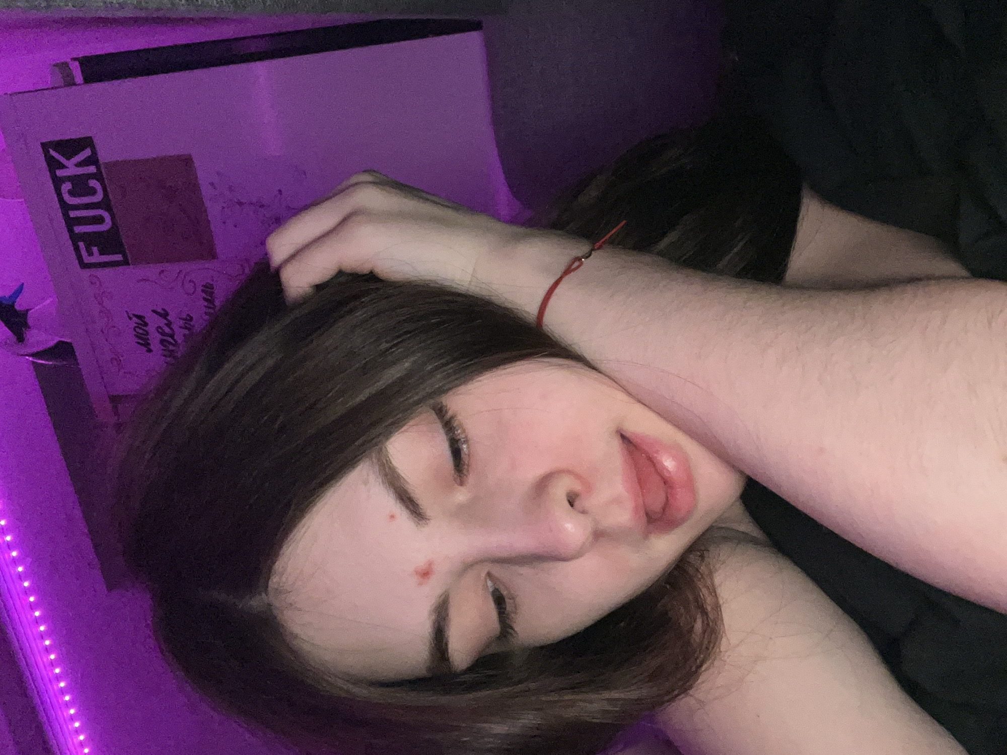 A young person with dark brown hair is depicted lying down indoors, likely in a bedroom, under dim purple ambient lighting. They are viewed from a high angle, with their eyes closed and mouth slightly ajar, revealing their tongue, suggesting a state of relaxation or sleep. One hand is gently placed on top of their head, with fingers intermingled in their hair. A thin red string bracelet is worn on their right wrist. A small red blemish is visible on their left cheek.
In the background, to the upper left, a light-colored box or book is partially visible. Prominently displayed on it is the English word "FUCK" in black letters within a black rectangular sticker. Below this, some Cyrillic text is faintly discernible, appearing to be "мои мечты" (my dreams) or similar, though it is largely illegible due to the angle and lighting. Along the far left edge of the frame, a strip of purple LED lights casts a soft, atmospheric glow. The overall atmosphere is intimate and quiet, possibly indicating evening or nighttime. This scene is set in Saint Petersburg, Russia.