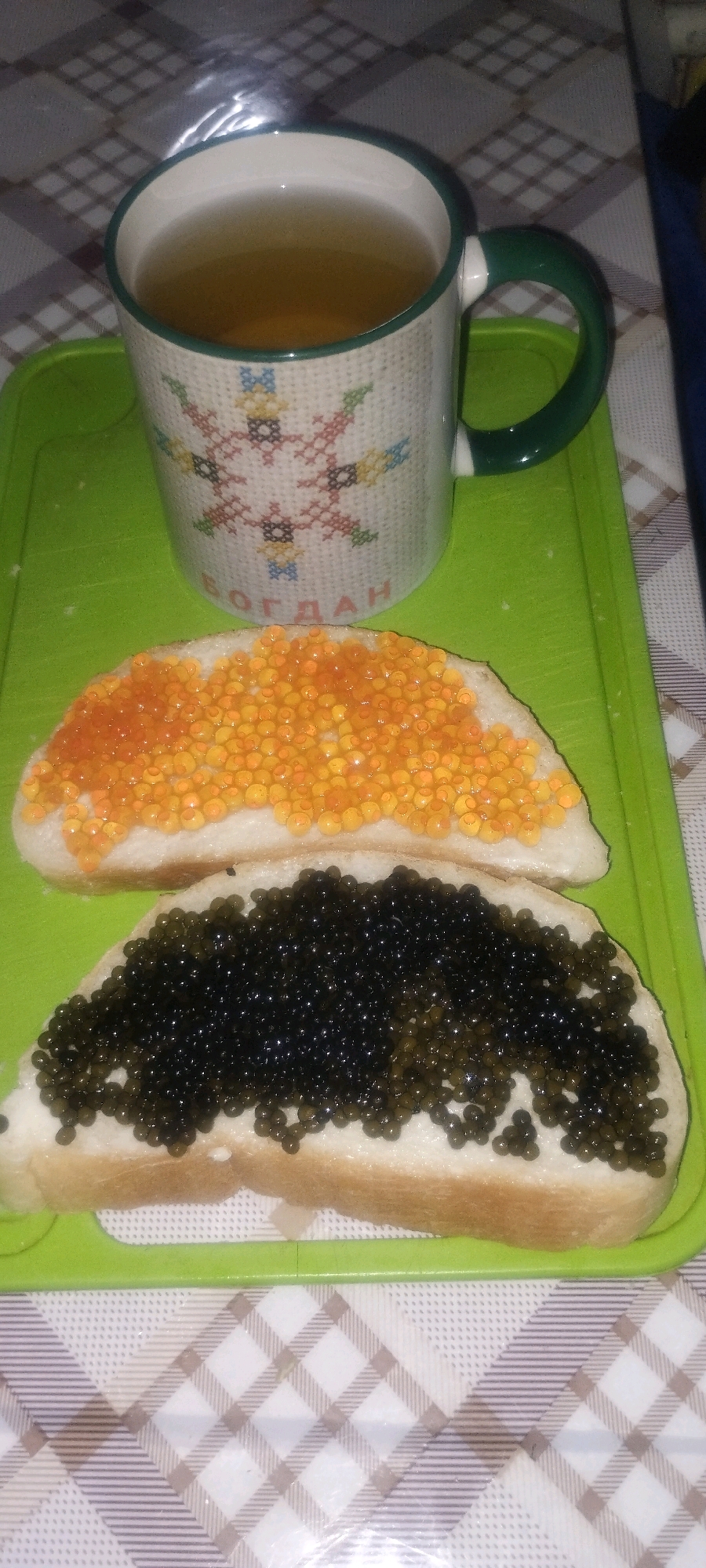 A close-up, overhead shot shows a green cutting board with a mug of tea and two slices of bread topped with caviar. The mug is white with a green handle and a pattern of embroidered-like figures and the Cyrillic word "БОГДАН" (Bogdan). The tea is a light amber color. One slice of bread is topped with bright orange caviar, and the other is topped with black caviar. The cutting board rests on a checkered tablecloth with shades of white, beige, and gray. The scene appears to be indoors, possibly in a kitchen or dining area. No specific location in Zaporizhzhia, Ukraine, can be determined from the image.