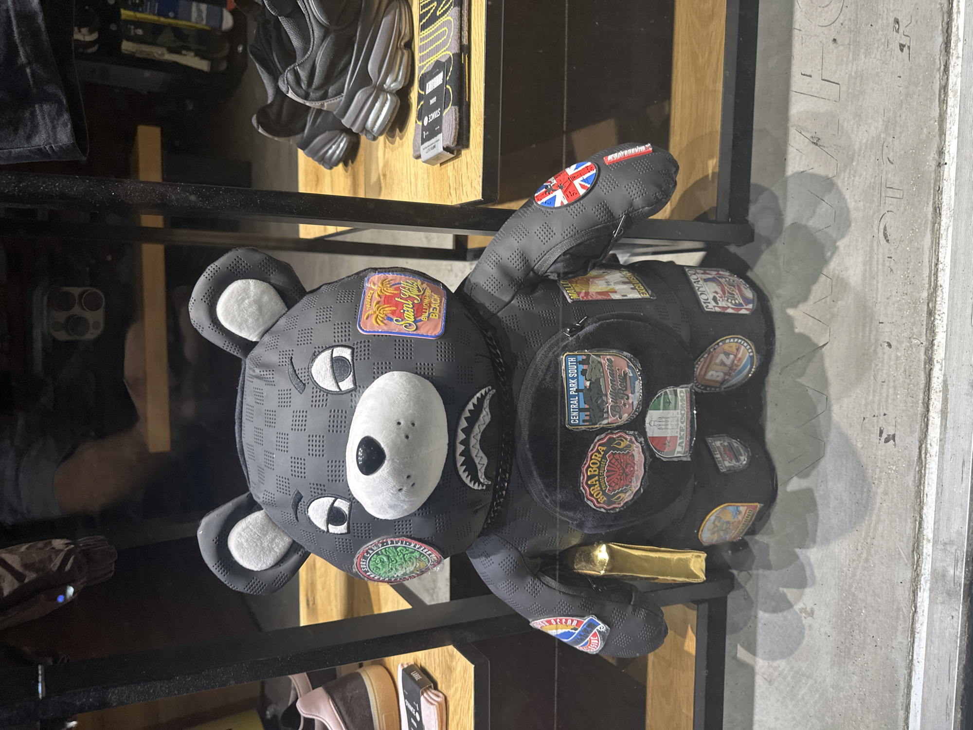 This media file captures a black, bear-shaped backpack or bag displayed in what appears to be a retail store in Sofia, Bulgaria. The main subject is the oversized, cartoonish bear bag, made of a black material with a perforated texture. Its face features a white snout, black nose, and stylized white and black eyes. A notable detail is the jagged, shark-like teeth design on the side of the bear's mouth.
The bag is adorned with numerous patches, suggesting travel destinations or luxury themes. Visible text on these patches includes "Saint Gold BILLIONAIRE BEACH," "PARADISE," a Union Jack flag, "CENTRAL PARK SOUTH," "MONACO," "BORA BORA Exclusive Cruises," "LAKE COMO," "IBIZA," and what appears to be "OCEAN SURF BREAK." Another circular patch displays text like "SURF CAMP WILD BREAKS."
The bear bag is positioned on a dark shelf, likely part of a display. In the background and around the bear, other retail products are visible, including grey athletic footwear on an upper shelf and a pair of socks with "STANICE" (Stance) text on them. A small tag with the word "LARGE" is also discernible. The floor below the display is concrete, showing reflections of objects and shadows, possibly from a phone or other items held by the person taking the photo. The overall scene suggests an indoor setting with artificial lighting, likely during store operating hours.