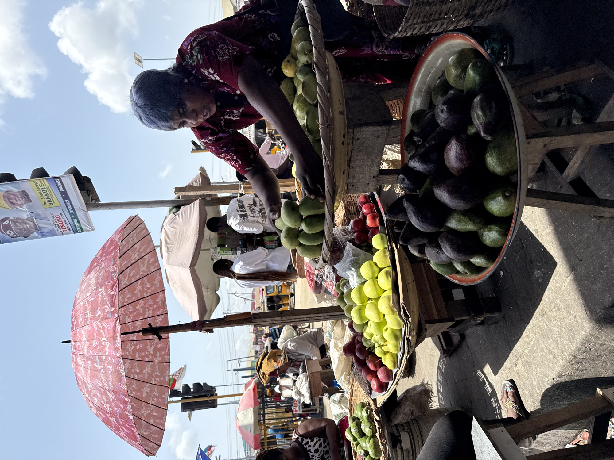 A woman with gray hair wearing a red floral shirt is selecting fruit from a basket at a roadside fruit stand in Ado, Nigeria. The stand displays a variety of fruits including avocados, green apples, and plums. An umbrella provides shade for the stand. In the background, other people, umbrellas, traffic lights, and buildings are visible. A political campaign poster is affixed to a nearby structure.