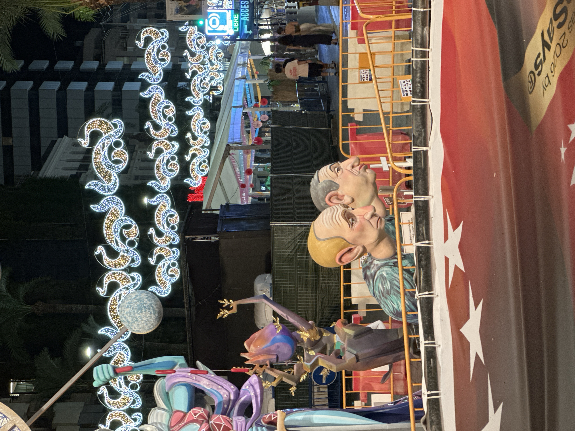 This image shows a street scene at night in Alicante, Spain, likely during the Fallas festival. The foreground is dominated by a large, colorful banner with white stars on a red and black background. In the middle ground, there are two large, cartoonish sculptures of human heads, one with blonde hair and the other with grey hair, positioned behind an orange safety barrier. Behind the sculptures, there are tents with lights and people walking around, as well as palm trees and buildings in the background. Decorative lights, forming elaborate swirling patterns, are strung across the street overhead. A green traffic sign with a symbol of a bicycle and the word "LIBRE ACCES" is visible on the right side of the image.