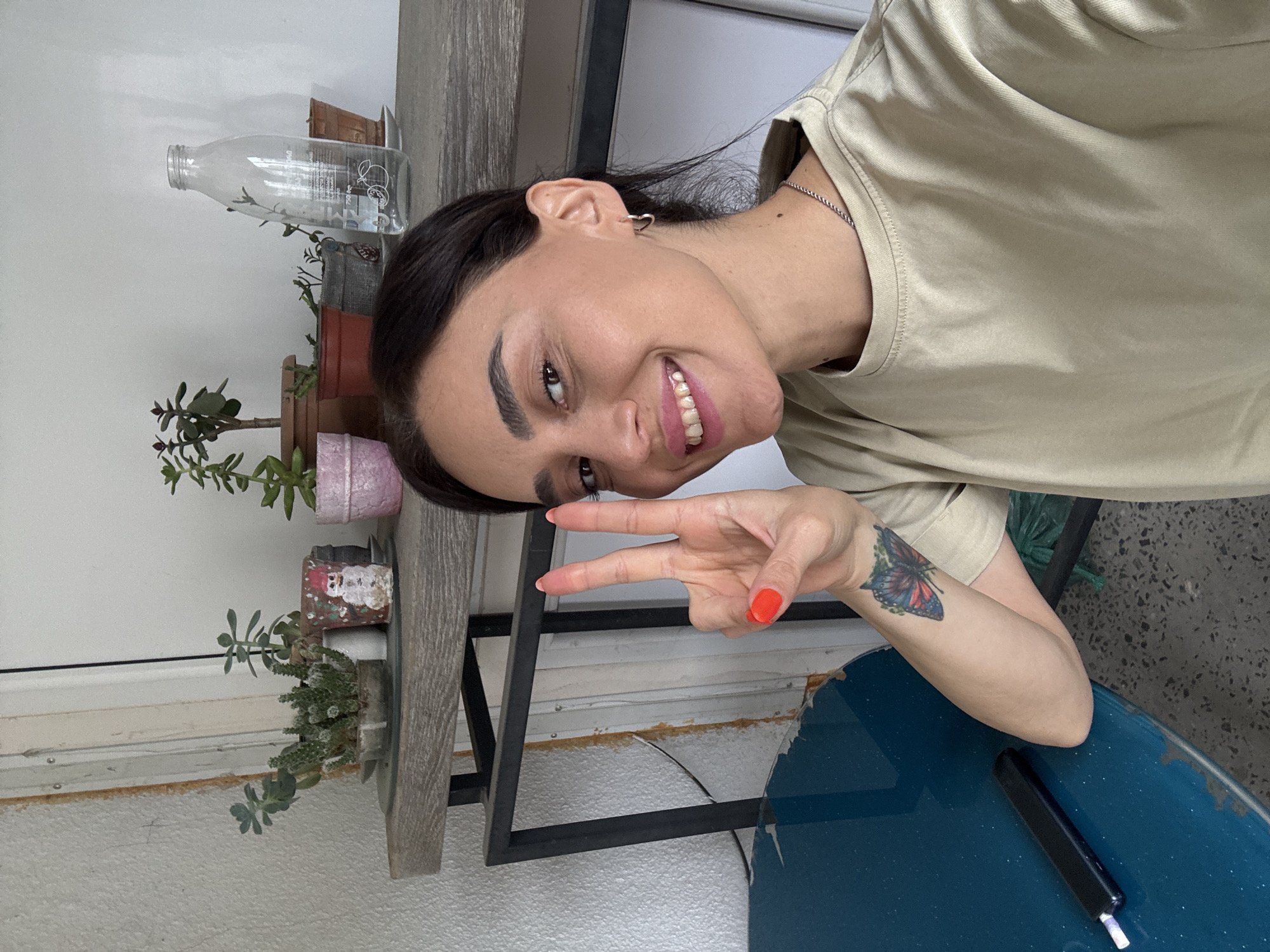 This media file is a portrait of a young woman in what appears to be an indoor setting with some plants and a shelf. The woman is smiling and making a peace sign with her hand. She has dark hair, dark eyebrows, and is wearing a light-colored t-shirt. On her left forearm, there is a tattoo of a butterfly. She has bright orange nail polish on her fingers.
The background includes a shelf with several potted plants, some of which are succulents. There is also a clear glass bottle with some liquid inside, and a dark blue surface in the foreground, possibly a table or desk. The lighting suggests it is daytime.
The image has been rotated 90 degrees counter-clockwise, so the woman is oriented sideways. The overall impression is casual and cheerful. There is no visible text in the image itself, other than what might be on the label of the glass bottle, which is illegible. The location context is Tbilisi, Georgia.