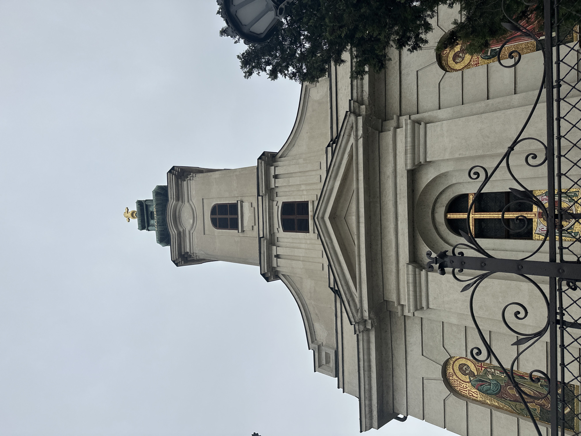 The image depicts an upward angle view of the exterior facade of The Name of Mary Church, located in Novi Sad, Serbia. The architecture is predominantly pale with a central tower that narrows upwards, capped with a gold cross. There are windows visible along the facade, some barred. The building is embellished with mosaic images. In the foreground, a wrought iron fence partially obscures the view of the church. Trees are interspersed around the building. The sky is overcast, giving the scene a diffused lighting.