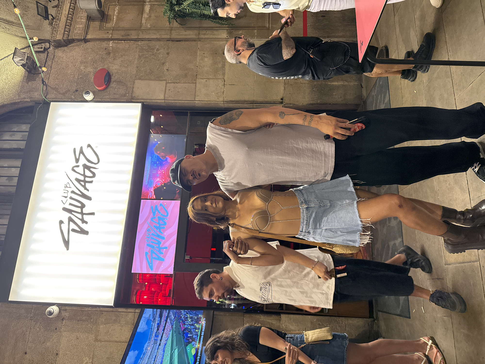 This is a medium shot of a group of people standing outside a venue in Barcelona, Spain. In the background, a large illuminated sign reads "CLUB SAUVAGE" in a stylized font. Below this, screens display the "CLUB SAUVAGE" logo and what appears to be a sports event. The venue has stone walls and a vaulted ceiling structure.
In the foreground, a woman with brown hair and a denim skirt is posing with her arm linked with a man wearing a grey sleeveless t-shirt and a black baseball cap. The woman is wearing a metallic-toned top with intricate detailing and dark brown boots. The man has tattoos on his arms.
To the left of the main couple, a woman with dark curly hair is wearing a black t-shirt and a denim skirt, and a man in a white t-shirt with dark shorts and sneakers. To the right of the main couple, two men are standing, one wearing a black t-shirt and the other a light yellow shirt. One of the men on the right is holding a phone and speaking to the man in the yellow shirt.
The ground is paved with stone tiles. There is a red rope barrier in front of a pink table on the right side of the frame. The overall atmosphere appears to be casual and social, possibly before or after an event.