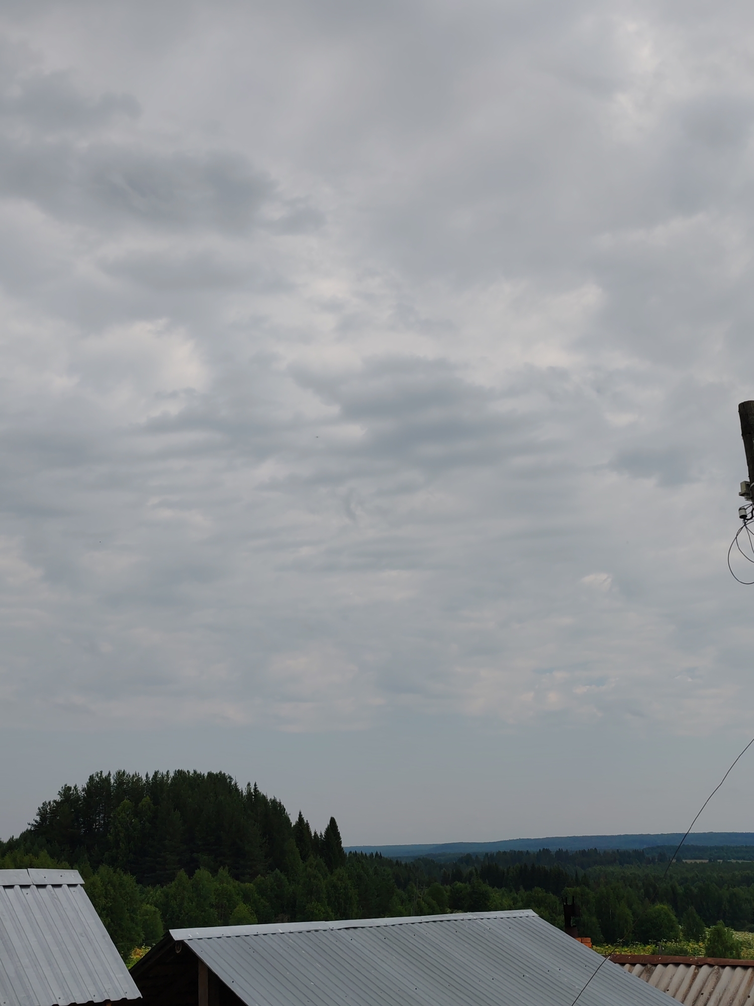 The image depicts an overcast sky above a rural landscape. In the foreground, the corrugated metal roofs of two buildings are visible. Beyond the buildings, a dense, dark green forest covers a hilly terrain. Further in the distance, layers of lighter green forests recede towards the horizon. The sky is filled with grey, textured clouds, with no clear sun visible. On the right side of the image, a dark, vertical pole with some wires and equipment attached is partially visible against the sky. The overall scene conveys a sense of a temperate, possibly humid, day in a natural, wooded environment. This scene is situated in the city of Chukhlem, Russia.