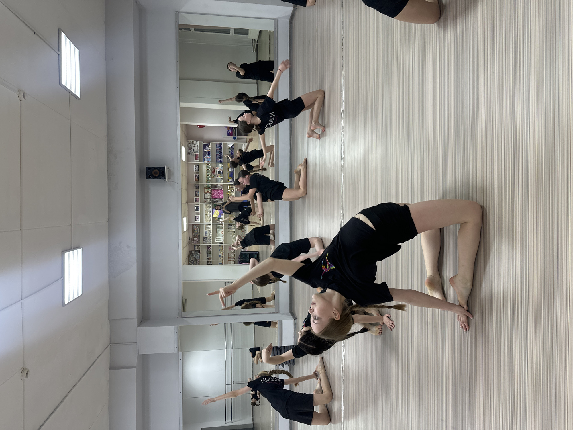 A dance class is in session in a studio in Votkinsk, Russia. Several young women wearing black shorts and t-shirts are performing a dance pose, bending backwards with one arm raised. The room has a light-colored wood floor, white walls, and large mirrors reflecting the dancers and the studio. A shelf filled with various items is visible behind the dancers in the mirror. Two rectangular lights are visible on the ceiling.