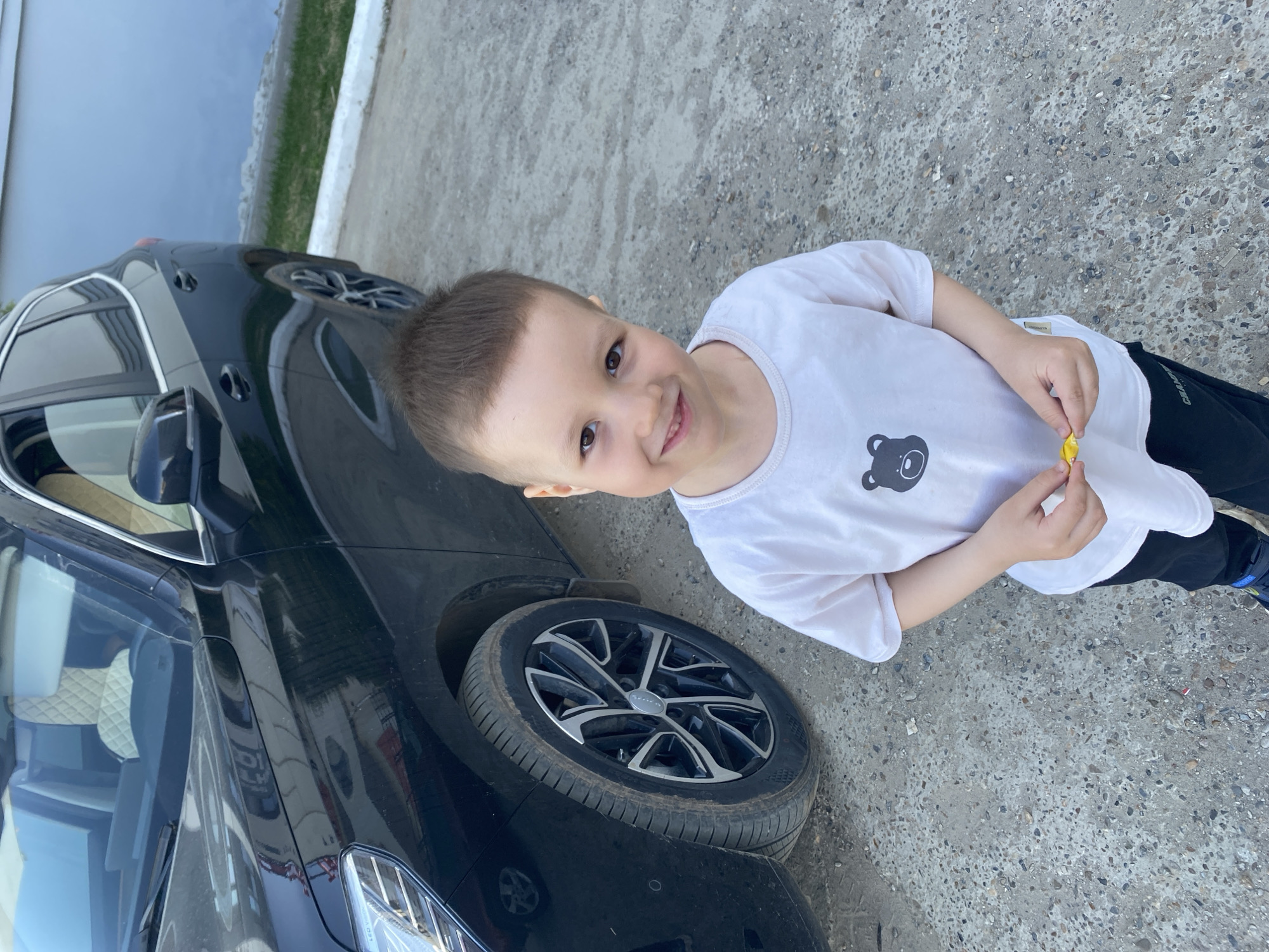 A young boy stands next to a black car parked on a concrete surface, possibly a sidewalk or parking area. The boy has short brown hair and is wearing a white t-shirt with a bear graphic and black pants. He holds a yellow object in his hands. The car is a dark color with silver and black rims. The image was taken during the daytime, with natural light illuminating the scene. There is no information about the image being taken in the city of Chaikovsky, Russia.