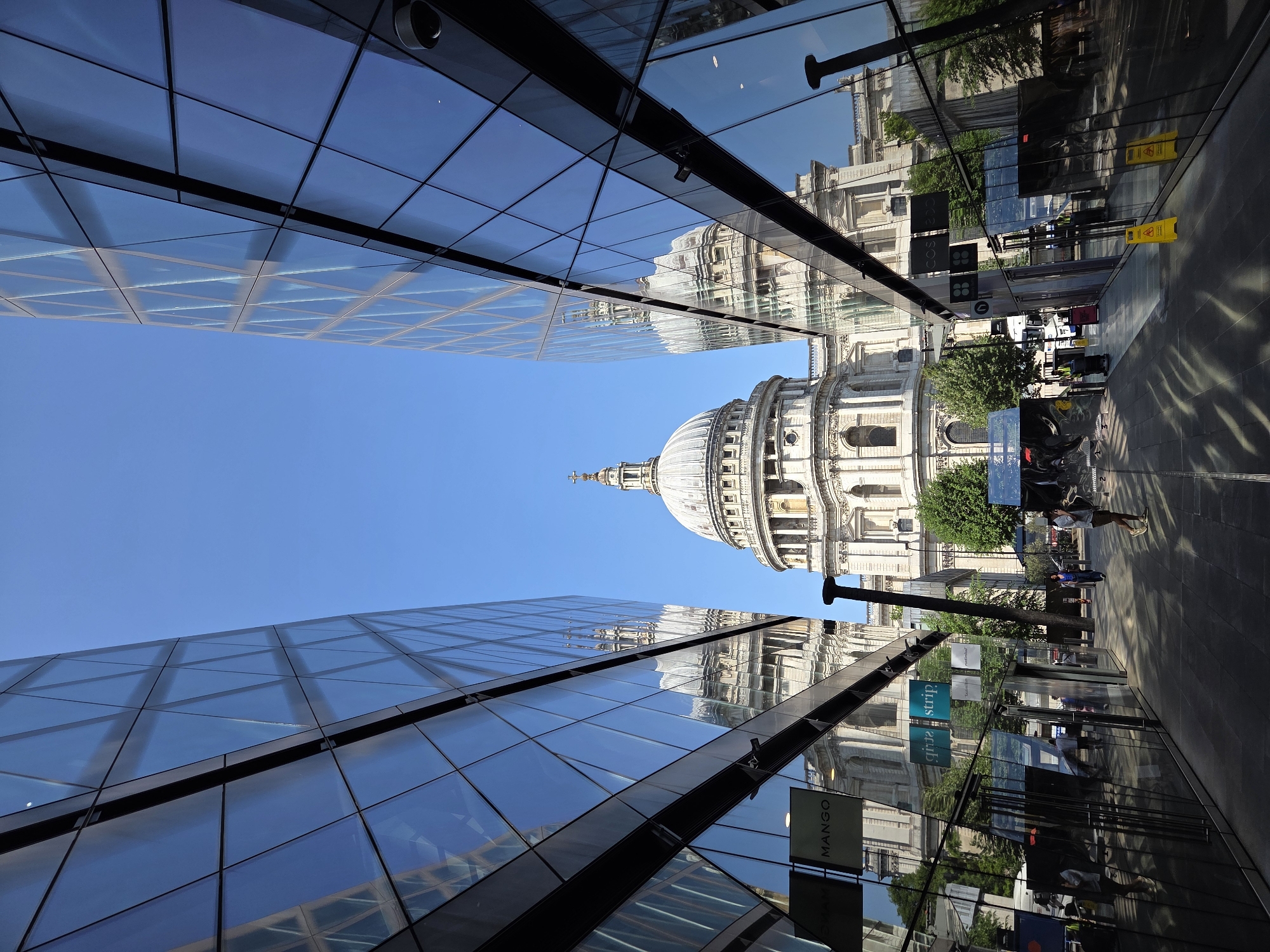 The image depicts St. Paul's Cathedral framed by modern glass buildings in London, United Kingdom. The cathedral's dome is visible in the distance, centered between the buildings. The glass buildings reflect the cathedral and the surrounding urban environment. Pedestrians are visible walking along the street. Street signs and store signs are also visible in the reflections. The sky is blue.