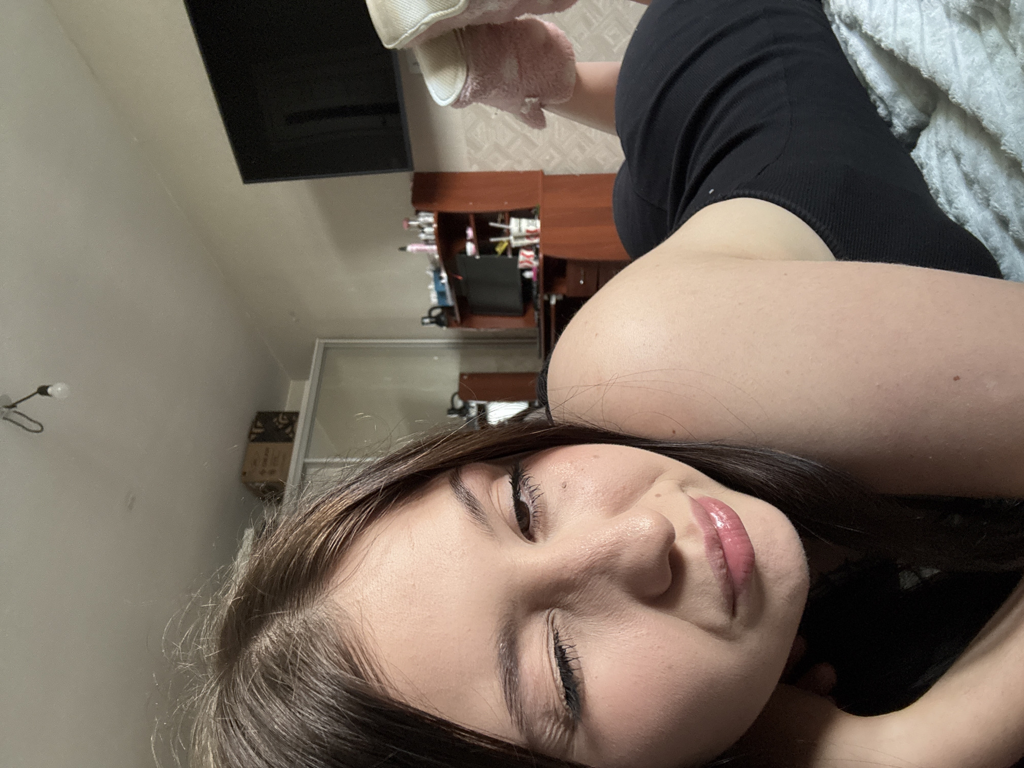 A young woman is lying on a bed, taking a selfie. She is wearing a black tank top and fluffy pink slippers. She has her right eye closed in a wink and her left eye is open. Her lips are slightly parted and glossy. The background shows a bedroom with a television, a dresser, and a closet. The image is taken from a low angle.