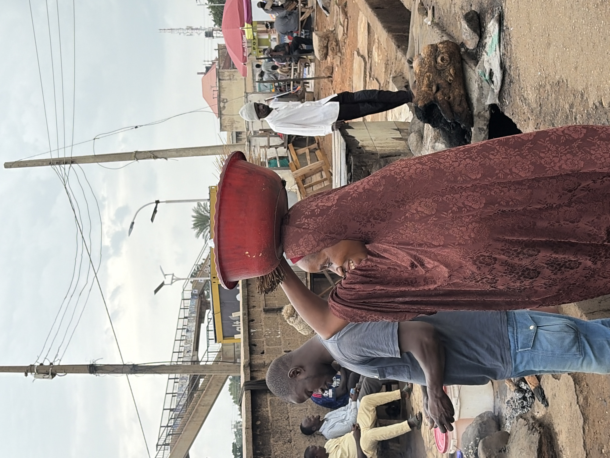 The image depicts a market scene in Gura Top, Nigeria. In the foreground, a woman is balancing a large red bowl on her head, covered with a patterned maroon shawl. A man stands next to her, seemingly assisting or interacting with her. Other individuals are seated in the background, engaged in various activities.
Overhead, a pedestrian bridge and power lines are visible. The environment appears to be a bustling marketplace with signs of commerce and daily life. The ground consists of a mixed surface including cracked concrete and dirt.