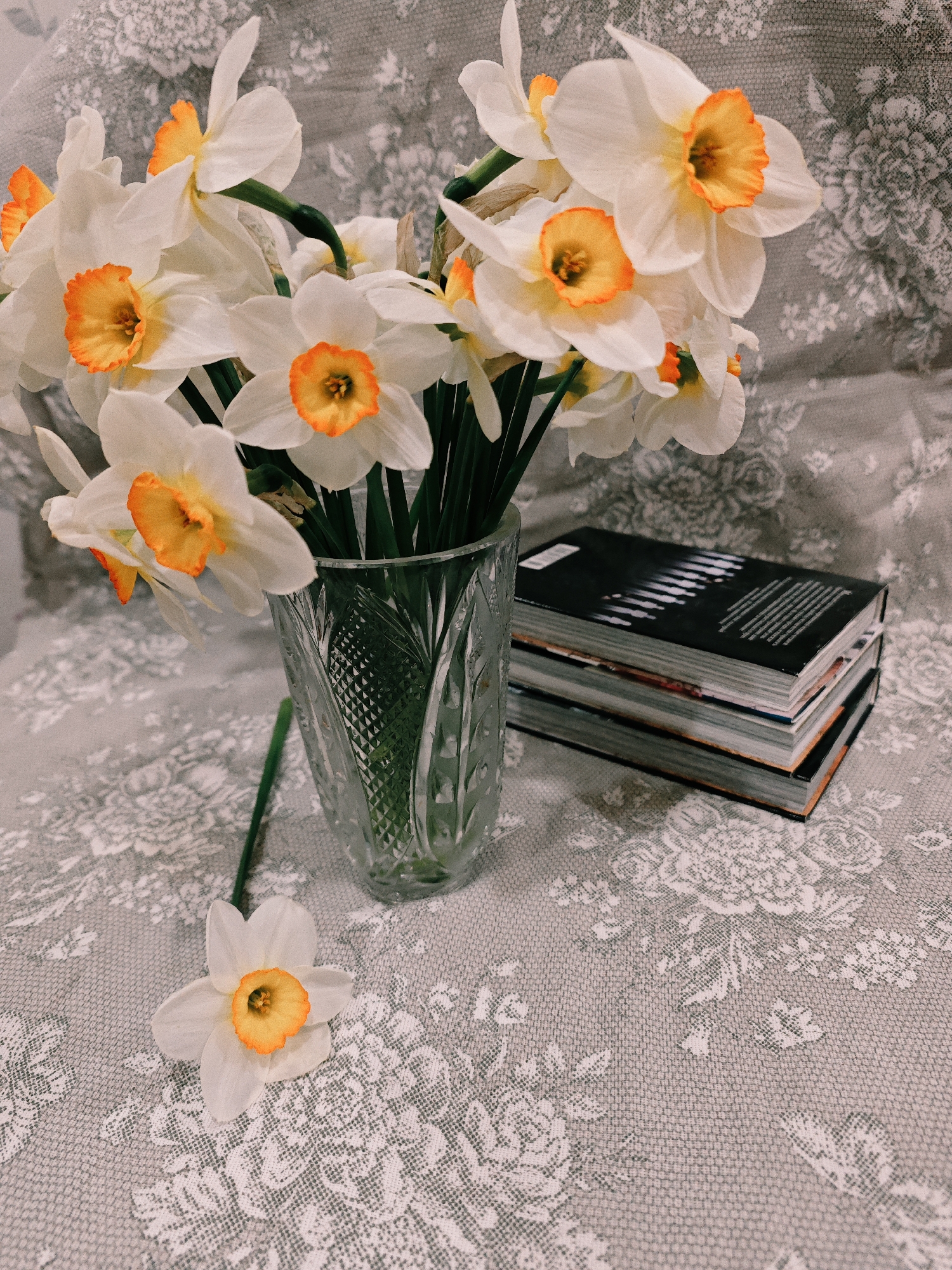 A still life features a bouquet of white and orange daffodils in a clear, cut-glass vase. A single daffodil lies next to the vase on a gray and white floral patterned surface. A stack of three books is placed to the right of the vase. The location is not discernible from the image, and there is no indication it is in the city of Markovskii, Russia. There are no people present.