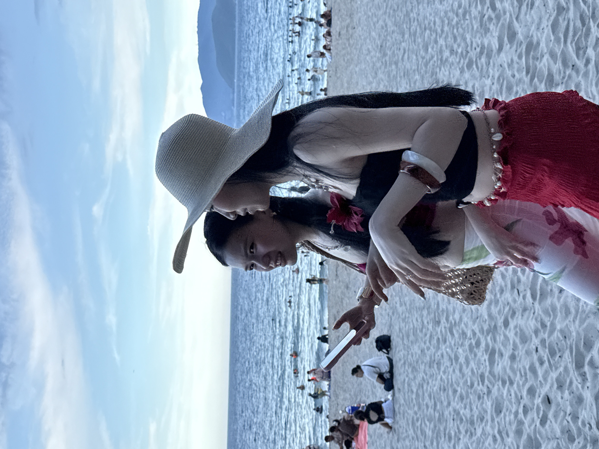The image shows two young women on a sandy beach, with the ocean and a distant mountain range in the background. The woman in the foreground is wearing a wide-brimmed straw hat, a black top, and a red, ruffled skirt. She is leaning in towards another woman, who is wearing a floral top and holding a smartphone. The second woman is looking at the phone, with her face illuminated by its screen. Several other people are visible on the beach and in the shallow water, some of whom appear to be swimming or sunbathing. The sky is partly cloudy, with a mixture of blue sky and white clouds. The overall scene suggests a casual day at the beach, possibly during the late afternoon or early evening, given the soft light. The presence of the ocean, sand, and people in swimwear points to a tropical or subtropical coastal location. The specific mention of Nha Trang Ward, Vietnam, provides a precise geographical context for this beach scene. No visible text is present in the image.