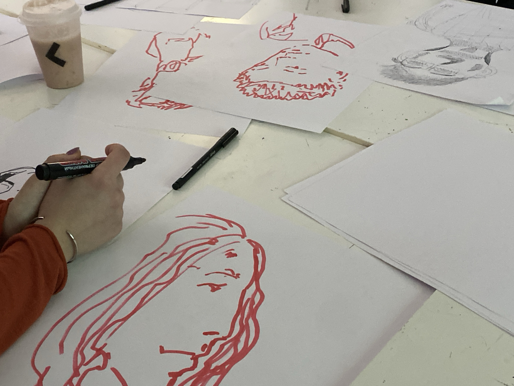 The image shows an indoor scene on a white table, likely in an art studio. A person wearing an orange long-sleeve shirt is holding a black marker and drawing a face outline in red on a piece of paper. There are several other sheets of paper scattered on the table, displaying various sketches in red and one in grey. A beverage in a plastic cup with a black logo is visible near the upper left corner. Another black marker lies on the table near the center. The environment suggests an artistic or creative workspace. No information in the image confirms the location is Moscow, Russia.