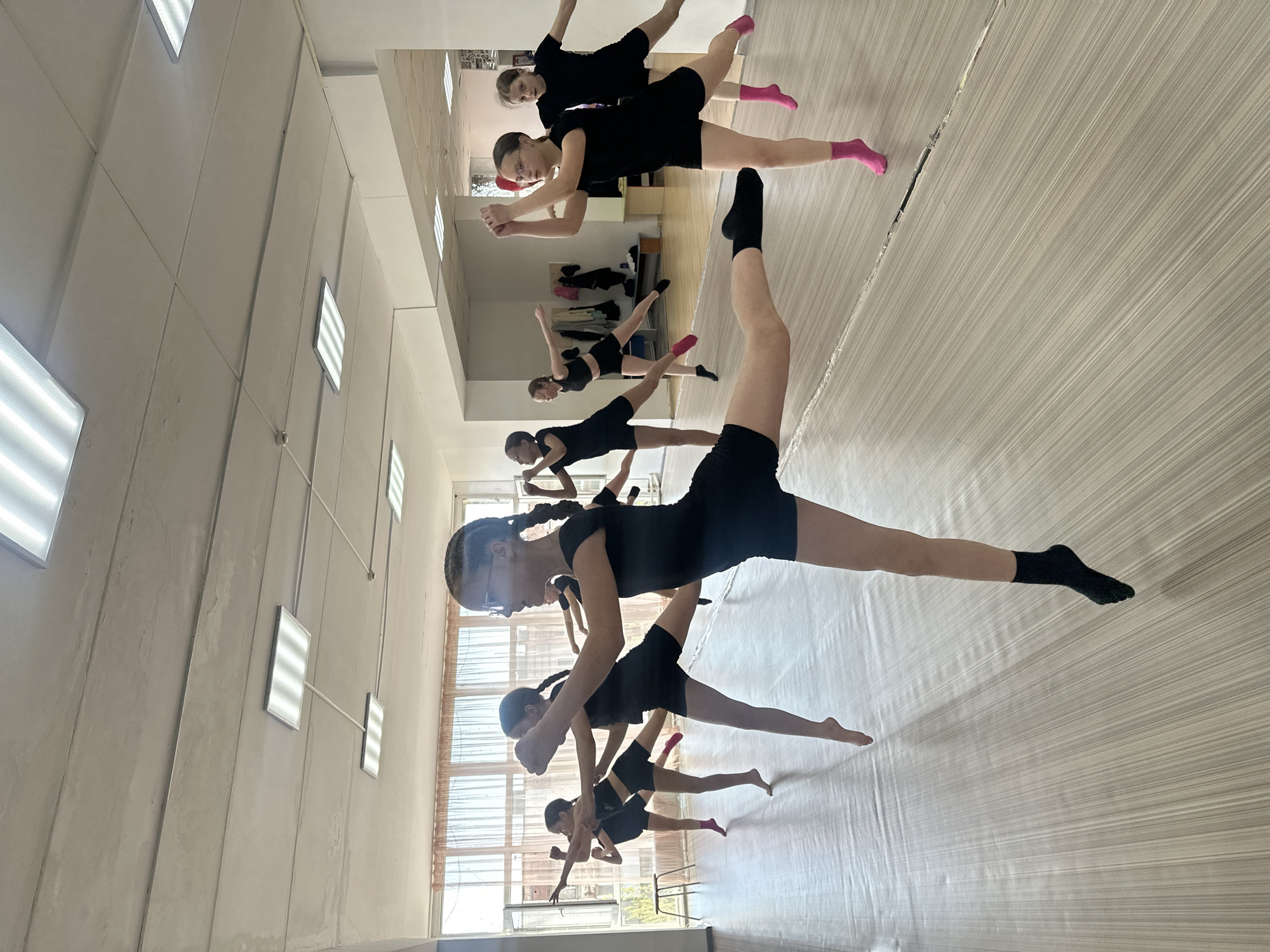 The image depicts a dance studio with several young dancers in black leotards and shorts. Some dancers are wearing socks, in hues of black and pink. The dance floor has a light-colored, lined surface. The dancers are engaged in a stretching exercise, with legs extended in a side split. The room has a mirrored wall reflecting the dancers and studio setup. Lighting fixtures are visible on the ceiling. Based on the availability of dance studios, the location is potentially Votkinsk, Russia.