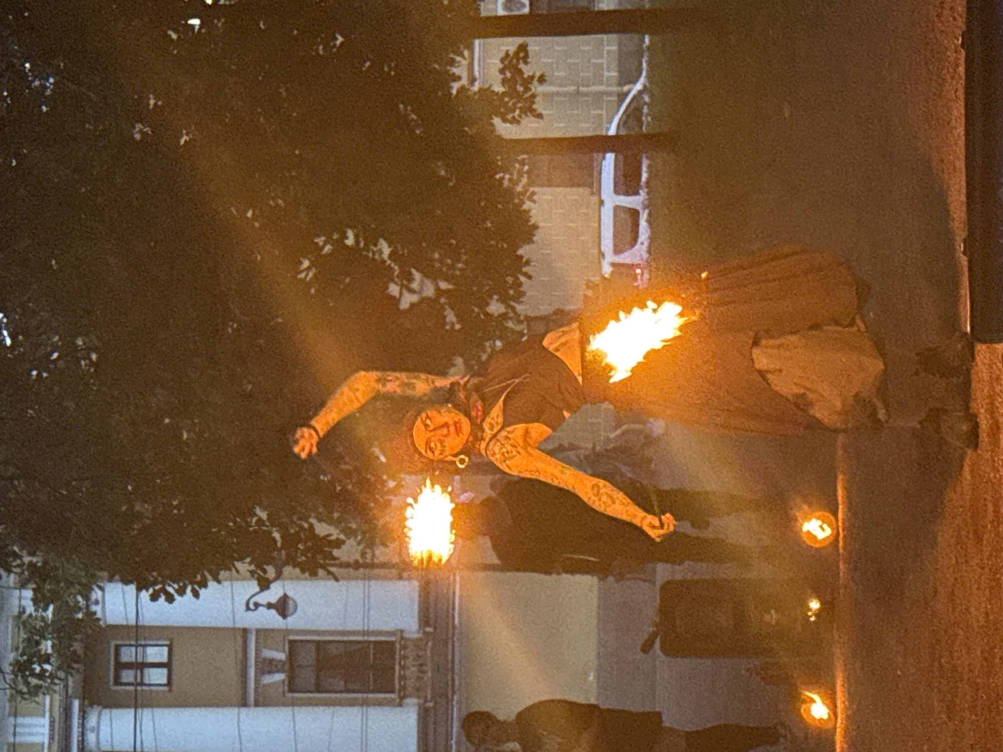 A fire performer is pictured in what appears to be Saint Petersburg, Russia, in front of an old building with classical architectural elements, including columns and arched windows. The performer, wearing a head covering and a dark vest over a light-colored skirt, is holding a fiery object above their head, creating a halo of flames. Another fire element is visible at their waist. Tattoos are visible on the performer's arms. In the background, other individuals are present, some partially obscured by smoke and fire. A white van is parked behind trees. Several small, flaming objects are scattered on the ground in the foreground. The scene is illuminated by the fire and appears to be taking place at dusk or in low light conditions.