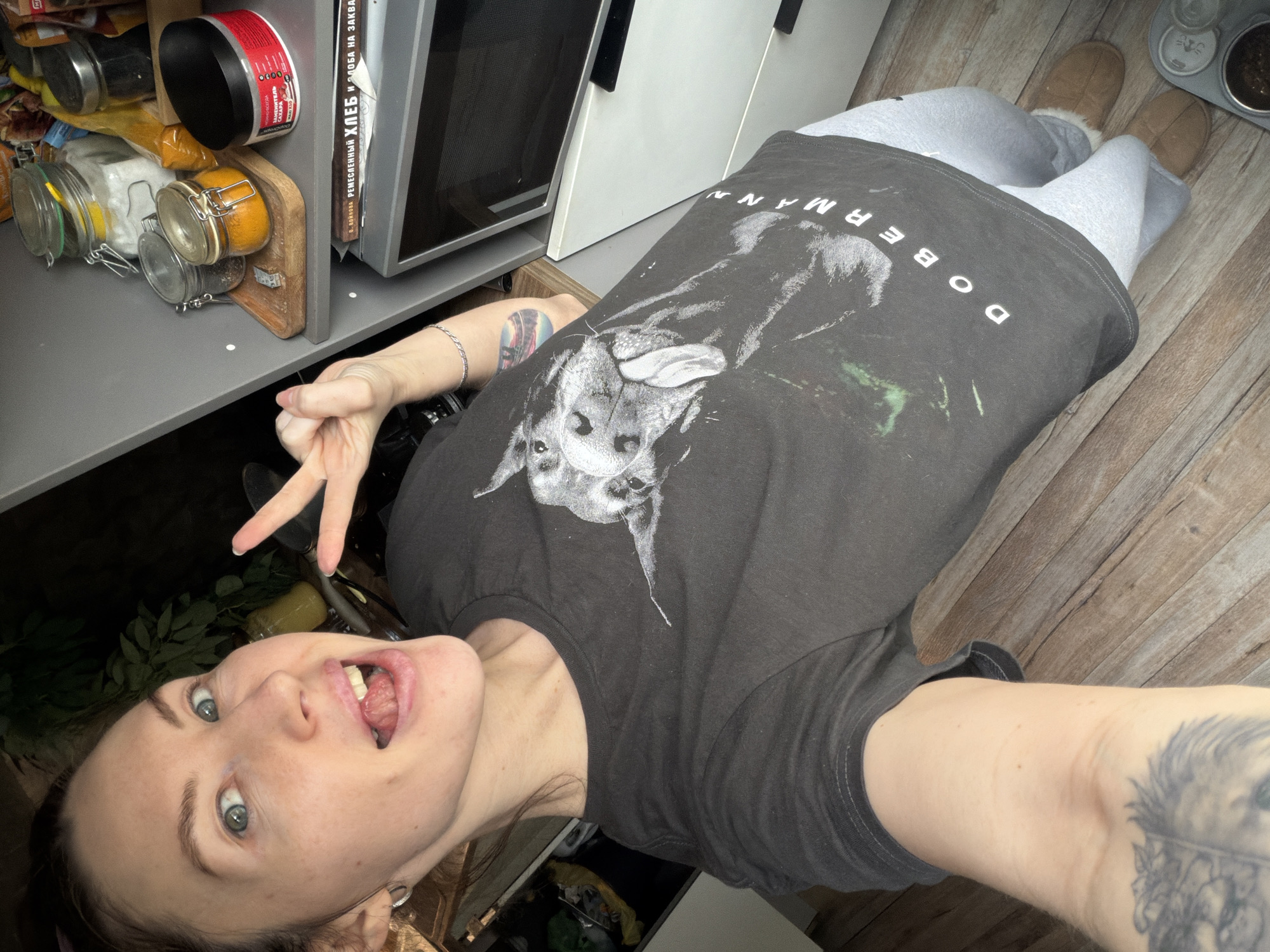 A selfie of a white woman with light eyes, displaying a playful expression with her mouth open and tongue visible, and giving a peace sign with her hand. She is wearing a black T-shirt with a Doberman dog graphic. She is also wearing gray sweatpants and brown slippers. The background includes a kitchen setting with a gray countertop cluttered with jars and containers, a white microwave, and a cookbook titled "РЕМЕСЛЕННЫЙ ХЛЕБ И ЗЛОБА НА ЗАКВ". The wooden floor suggests an indoor location. The presence of a cookbook written in Russian, titled "Войкова", could possibly indicate that the location of the photo is in Perm, Russia.