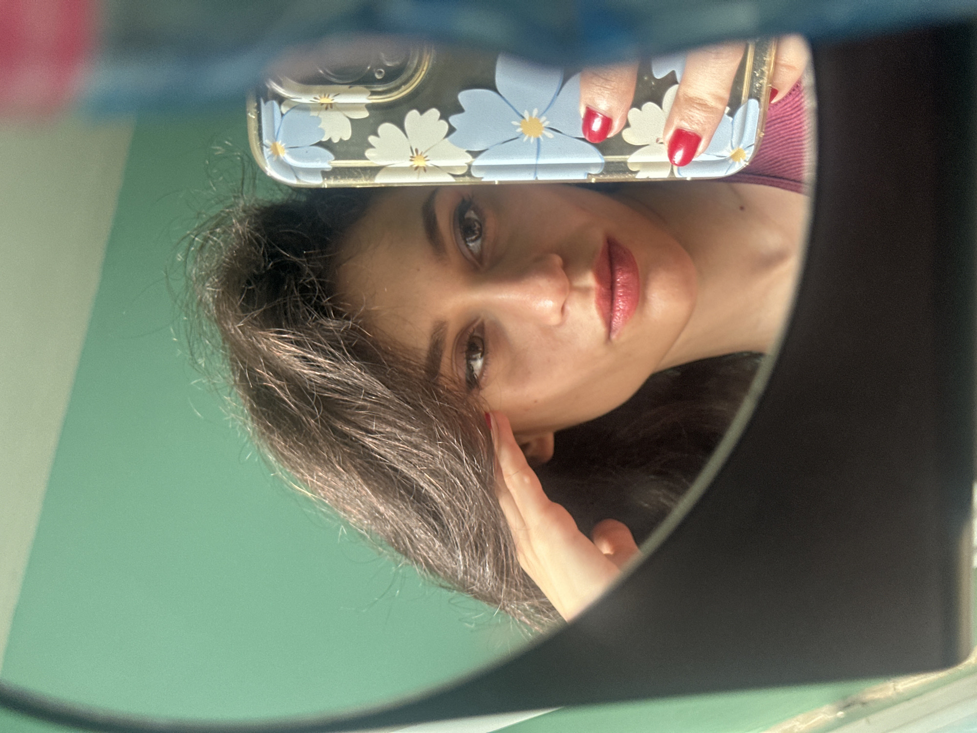 The image is a selfie taken by a young woman with dark hair and light skin, reflected in a mirror. She is wearing a pinkish top and has red nail polish. She is holding her right hand to her forehead, as if shielding her eyes from the sun or adjusting her hair. The background behind her is a teal green wall. A phone with a floral case is visible in her hand, also reflected in the mirror. The lighting suggests it is daytime. The image does not contain any information about the location in Sofia, Bulgaria.