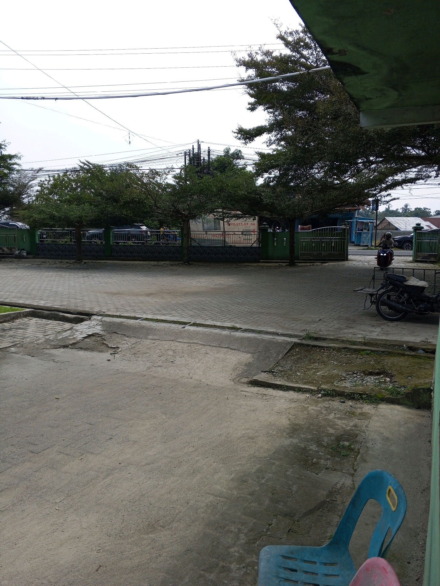 The image shows a street scene in Deli Tua Timur, Indonesia. A blue plastic chair is in the foreground, with a pink object partially visible to its right. Behind the chair is a paved area with some patches of discolored concrete and scattered debris. Further back, a gray brick pathway is visible, bordered by a concrete curb. Beyond the pathway is a paved area with a dark-colored motorcycle parked on the right side. The motorcycle has a sidecar attached. In the mid-ground, there's a green metal gate and a fence. Behind the fence, several parked vehicles and trees are visible. A person is riding a motorcycle in the distance, moving away from the camera. The sky is overcast, and there are power lines suspended across the scene. The overall impression is of an outdoor public space, possibly near a school or other institution, given the presence of a fence and gate.
