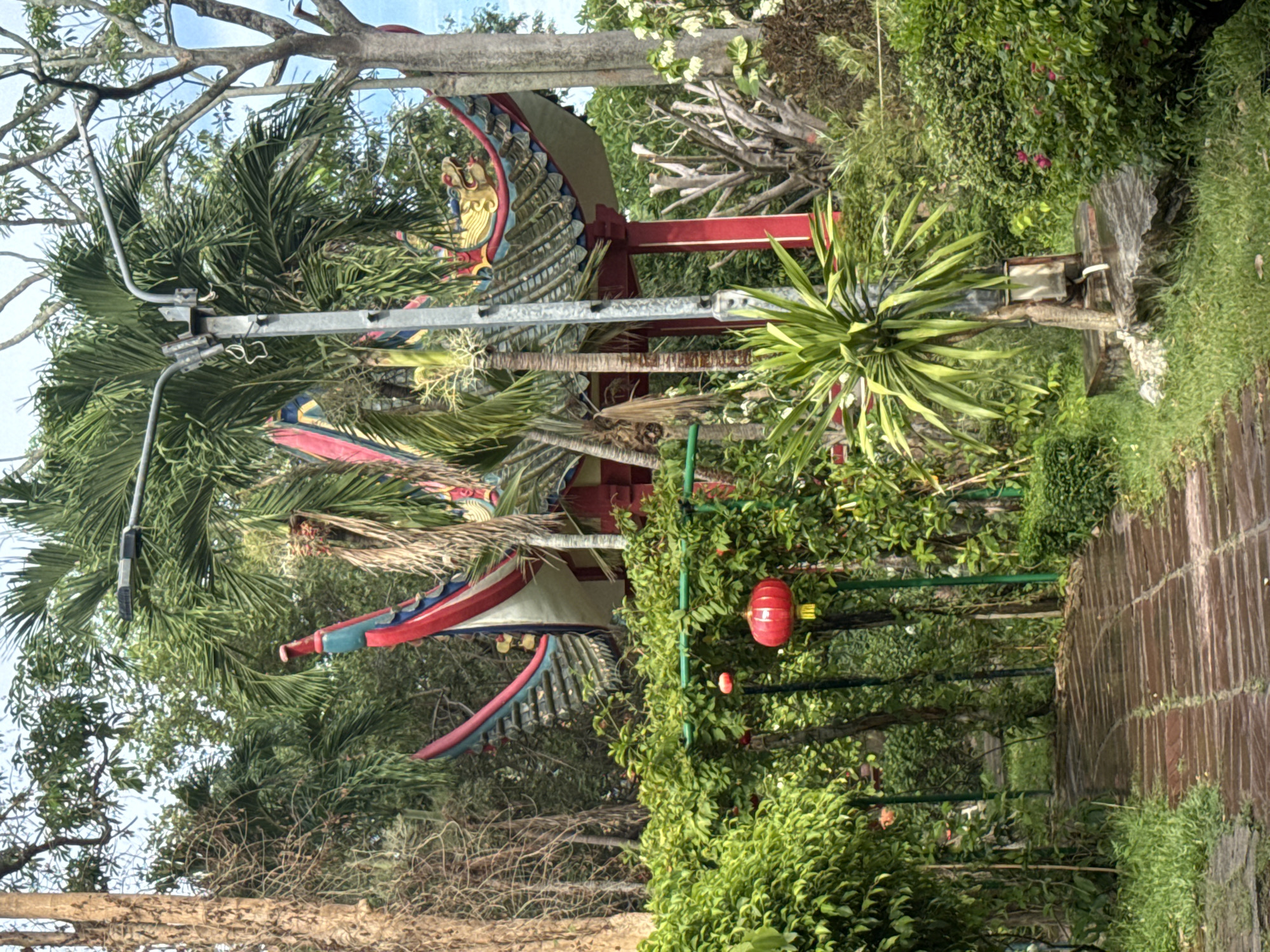 A sideways, eye-level shot depicts a landscape in มาบฟักทอง, Thailand, featuring a tiled walkway on the right that curves into the scene. Dense greenery and bushes line the path, with red lanterns hanging among the leaves. A traditional Thai structure is visible, its roof adorned with red, blue, and yellow ornamentation, including dragon motifs. The structure is supported by red columns. Palm trees are interspersed throughout the scene, adding to the tropical atmosphere. A silver metal post is visible near the Thai structure. The background includes additional trees and a hint of the sky. The scene is rich in natural elements and cultural details.