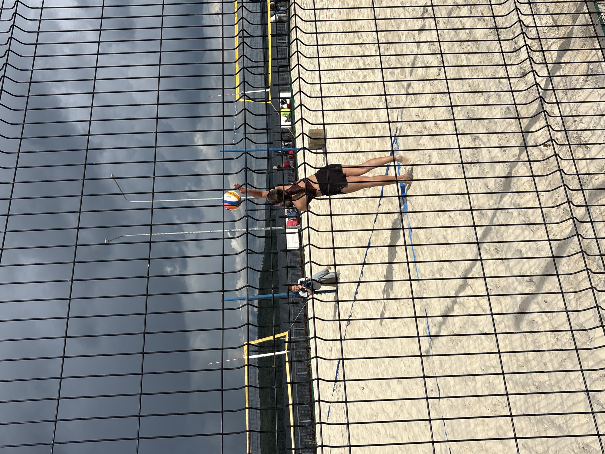 The image depicts a beach volleyball court on a cloudy day, viewed through a wire fence. In the foreground, the fence creates a grid-like overlay on the scene. On the court, a young woman in a dark red sports bra and black shorts is in the process of serving a volleyball, her arm extended upwards. To her left, a person in a white t-shirt and grey pants is sitting, possibly watching the game. The court is made of sand, with blue lines marking the boundaries. In the background, a volleyball net is visible, along with trees and a body of water. The sky is dark with storm clouds. This scene is likely located at the beach volleyball courts near the Kama River embankment in Chaikovsky, Russia.