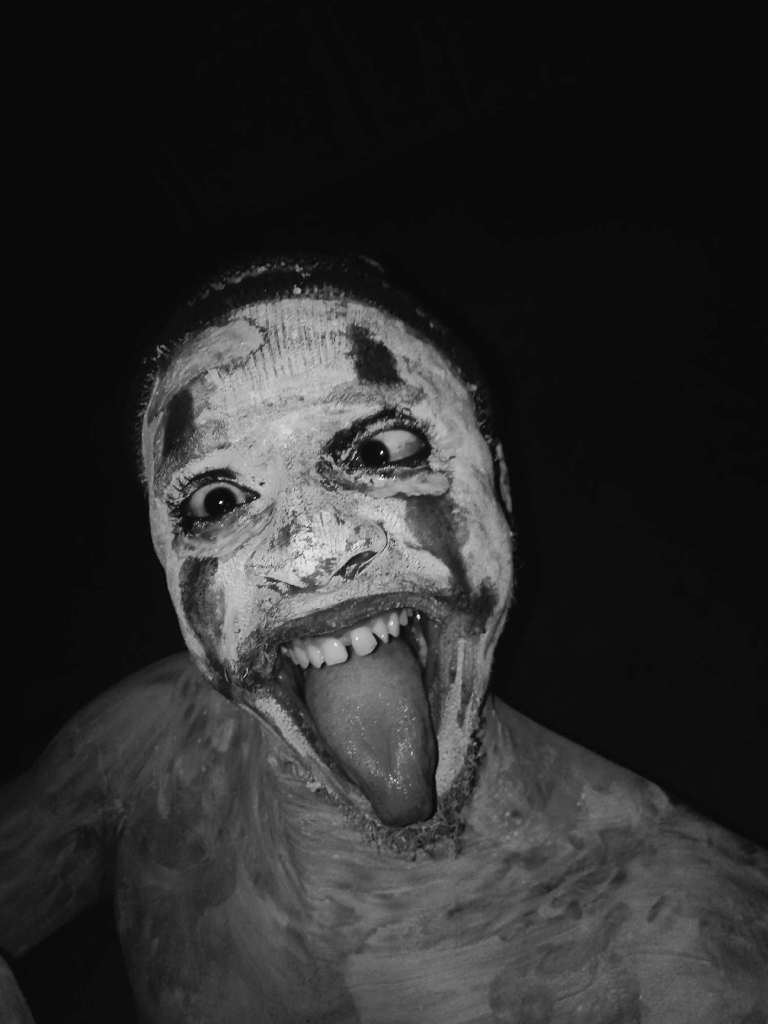 A black and white, close-up, low-angle shot captures a person with a distorted facial expression and body covered in white paint. The person's eyes are misaligned, and their mouth is wide open, revealing teeth and a protruding tongue. The background is a solid black. The location is unknown.