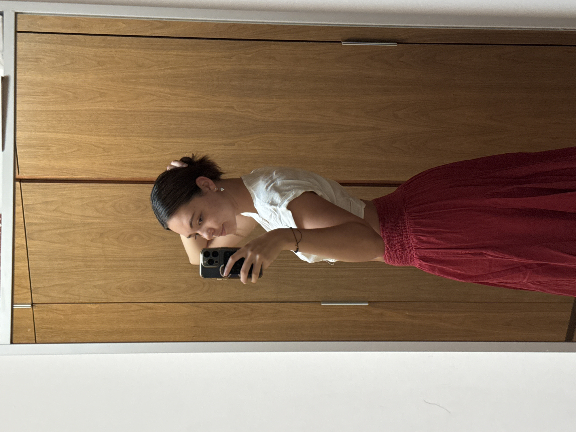A young woman is taking a selfie in a mirror. She is wearing a white top and a red skirt. She is holding a black smartphone. The background appears to be wooden closet doors. There is a white border around the mirror. The location in the city of San Bernardino Tlaxcalancingo, Mexico, cannot be determined from the image.