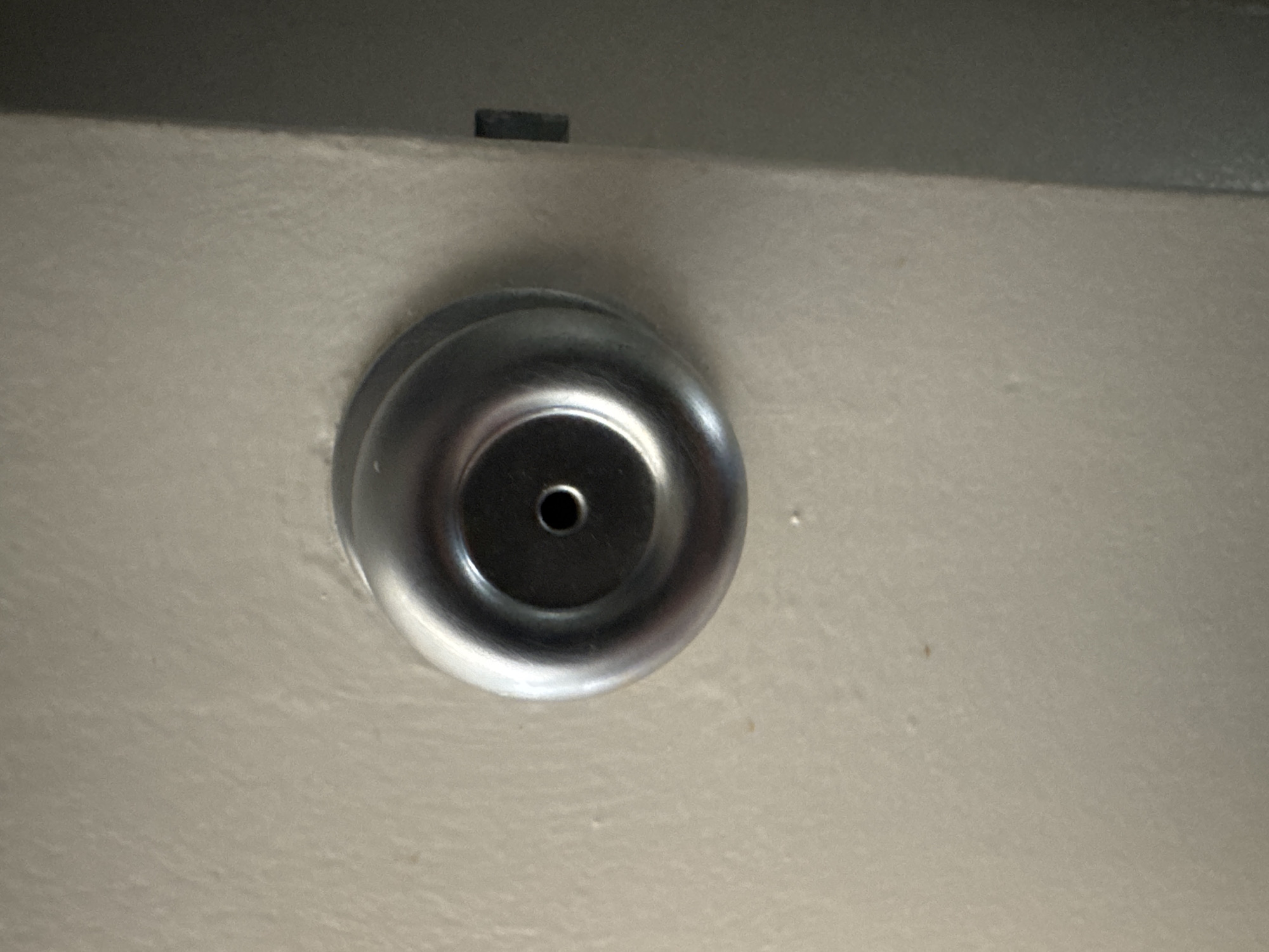A close-up, slightly angled shot shows a silver, round doorknob mounted on a light beige, textured wall. The doorknob has a circular indentation in its center with a small, dark hole. The wall shows some minor imperfections, including a few small, light brown specks. To the right of the doorknob, a dark vertical line suggests the edge of a doorframe or a seam in the wall. The lighting casts a slight shadow on the right side of the image, indicating a light source from the left. The scene appears to be indoors, likely in a residential or commercial building, but its specific location within Los Angeles, United States of America, cannot be determined from the visual information provided.