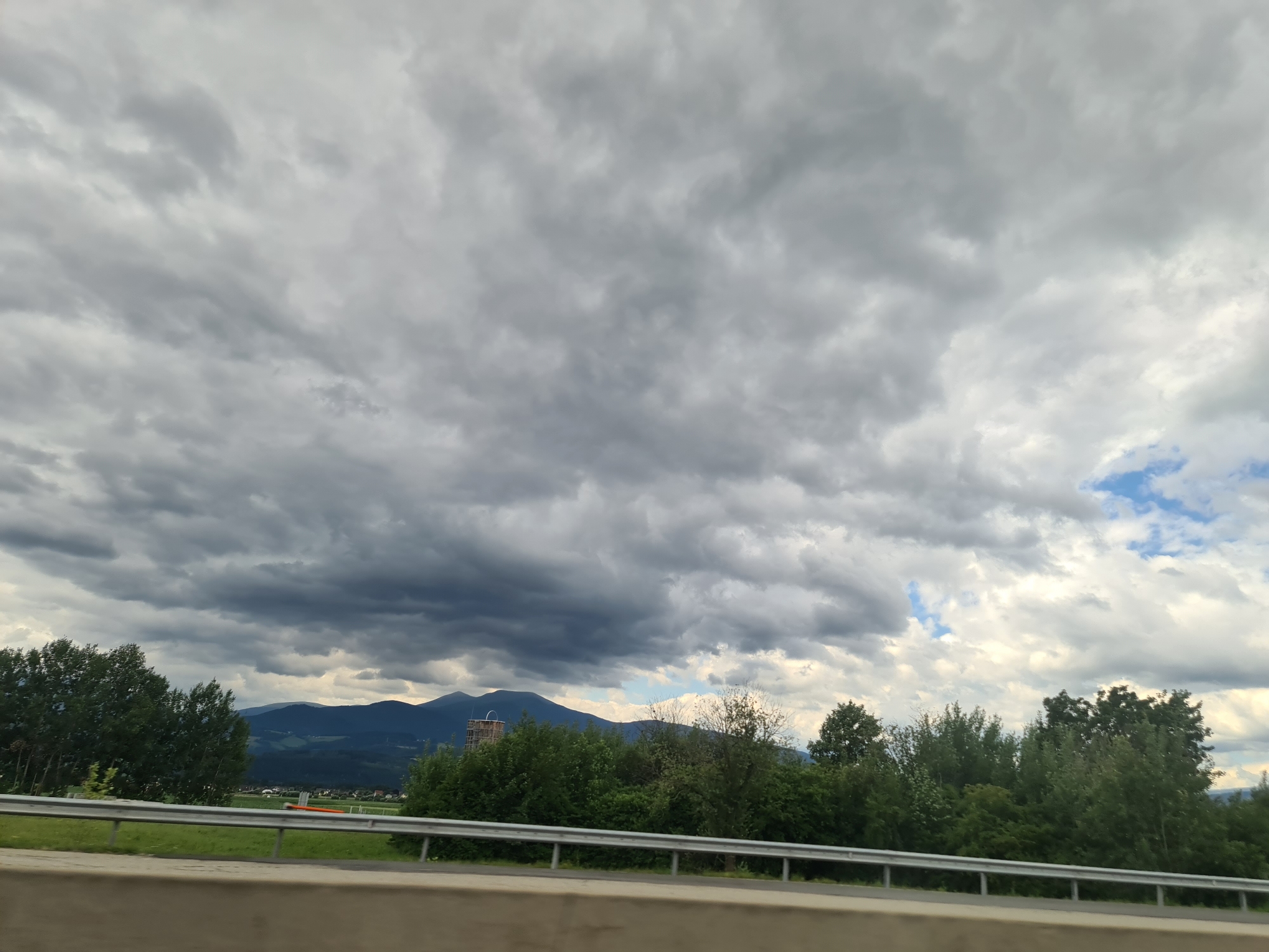 The image depicts a landscape scene, likely captured from a moving vehicle, with a focus on the sky and distant mountains. The sky is heavily clouded with a mix of dark and light gray formations, suggesting an overcast or impending stormy weather. Patches of blue sky are visible through breaks in the clouds on the right side of the frame.
In the middle ground, a line of green trees forms a dense border, with varied tree heights and foliage. Behind the trees, a mountain range with distinct peaks is visible, appearing distant and somewhat hazy. A notable structure, resembling a partially constructed tower or industrial building, stands out against the trees and mountains on the left side of the frame.
The foreground shows a guardrail, characteristic of a highway or road, with a strip of green grass and a light-colored barrier below it. This suggests the photo was taken from a roadside vantage point, possibly a car.
The overall impression is one of a natural landscape under a dramatic sky, with human-made elements present but not dominating the scene. The specific location in Pausendorf, Austria is not visually identifiable within the image itself.