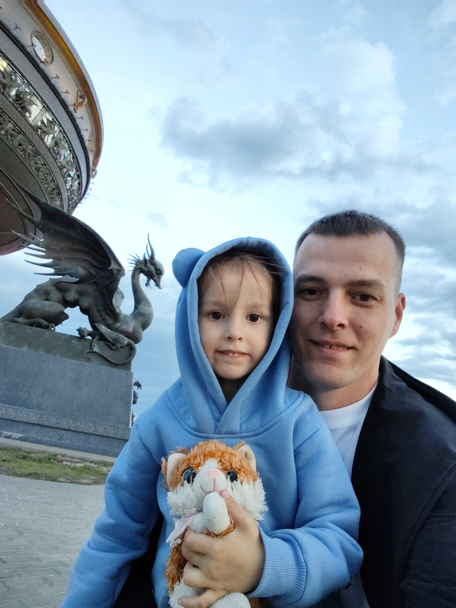 A portrait-style photograph captures a man and a young child posing outdoors against a cloudy sky. The man, on the right, has short dark hair and a clean-shaven face, smiling directly at the camera. He wears a dark jacket over a white t-shirt. The child, positioned in front of him and to the left, also smiles at the camera. The child is wearing a light blue hooded sweatshirt with small, rounded ears on the hood, and holds a small, fluffy orange and white stuffed animal, resembling a fox or cat, with both hands.
In the background, to the left, a large, ornate circular building with intricate metalwork is visible. Adjacent to this structure stands a prominent bronze statue of a winged dragon with scales and horns, resting on a stone base. This dragon statue is characteristic of Zilant, a mythological symbol of Kazan. The sky is overcast with visible clouds, suggesting a cloudy day. The ground in the foreground appears to be paved. The subjects appear cheerful and relaxed, likely at the Kazan Family Center (Kazan Wedding Palace) in Kazan, Russia. No visible text is present in the image.