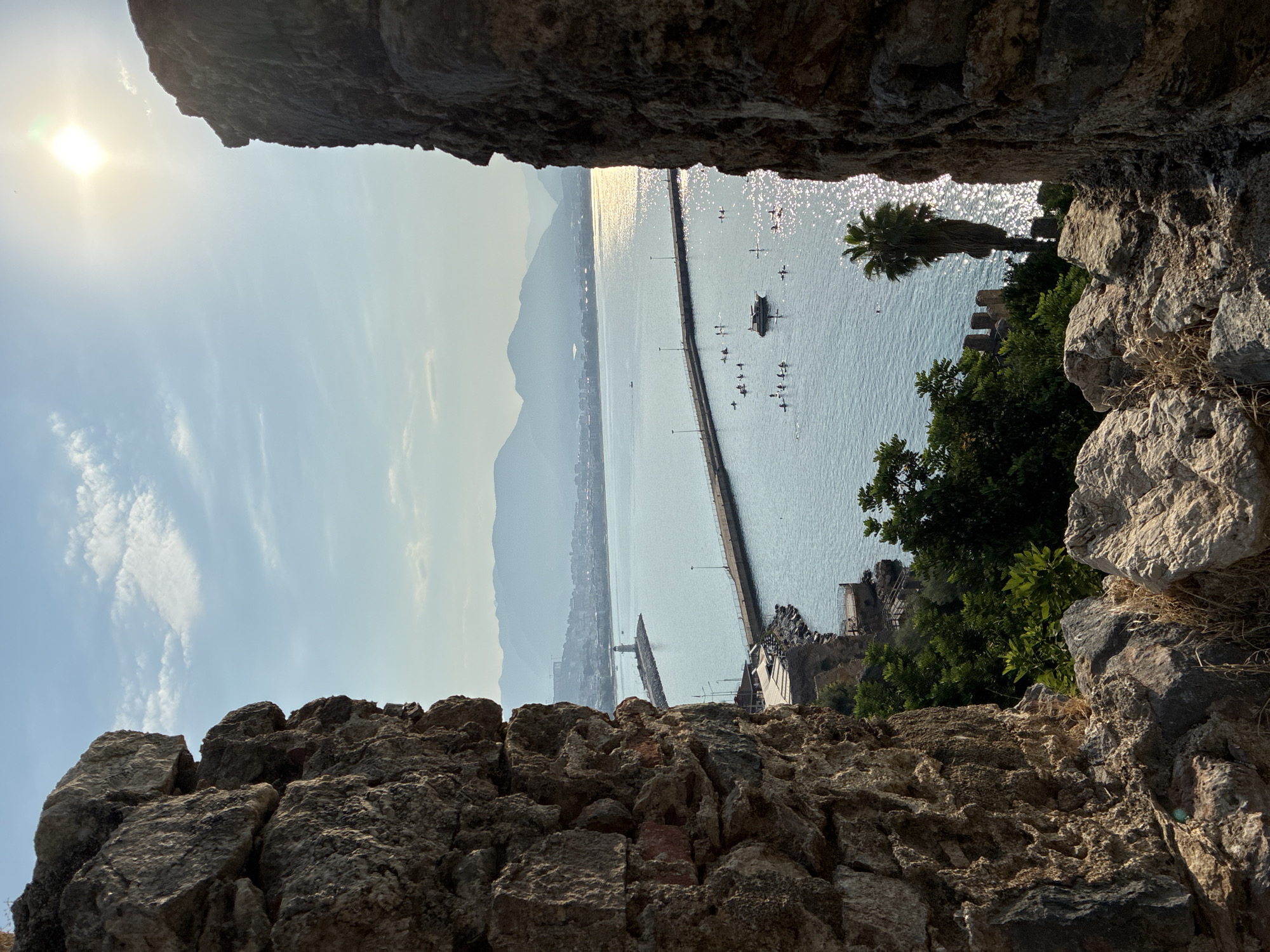 The photograph captures a view through a stone archway overlooking the Mediterranean Sea in Alanya, Turkey. In the foreground, weathered stone walls form the frame of the scene. Beyond the archway, a wide expanse of the sea glitters under a bright, hazy sun. A long pier with lampposts extends into the water, leading to a lighthouse on its end. Numerous people are scattered across the water, some on paddleboards and others possibly swimming or engaged in other water activities. A small boat is also visible among them. The coastline, dotted with buildings and lush greenery, stretches into the distance, with hazy mountains forming the backdrop. A solitary palm tree stands on the right side of the frame, its silhouette partially obscured by the sun's glare. The overall impression is of a serene, sun-drenched coastal landscape.