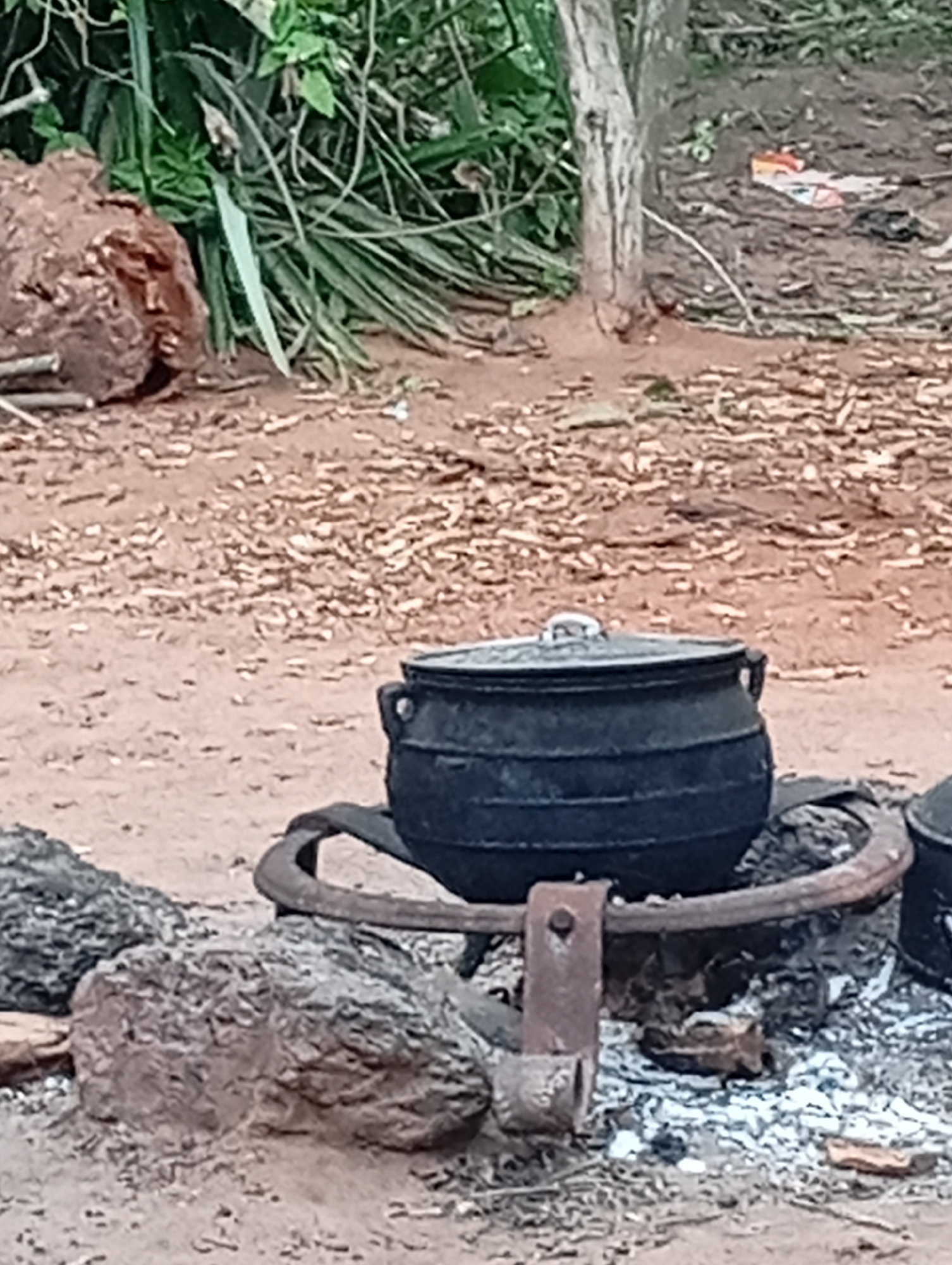 The image depicts an outdoor scene likely in Adda, Nigeria, focusing on a cooking setup. A large, black cast-iron pot with a lid is placed on a rustic metal stand over a fire pit with smoldering embers. The pot appears to be of significant size, suitable for communal cooking. The stand is constructed from what appears to be repurposed metal, with a U-shaped bracket supporting the pot's rim. The surrounding ground is composed of reddish-brown earth and scattered small stones and debris, indicative of a natural or semi-developed environment. In the background, lush green vegetation and a tree trunk are visible, suggesting a rural or semi-rural setting. A large, weathered piece of wood or bark lies to the left of the frame, and some discarded items, possibly packaging, are scattered in the background. The overall impression is one of traditional cooking methods being employed in an informal outdoor setting. There are no people visible in the image.