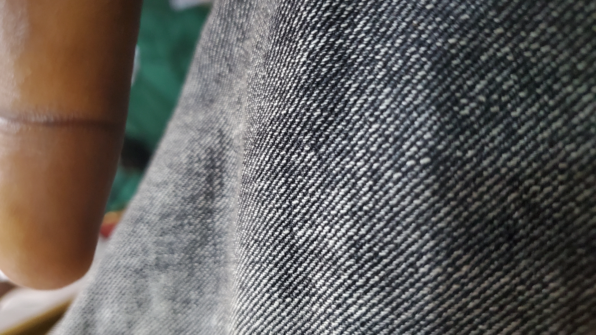 A close-up, macro shot focuses on the texture of dark denim fabric. The weave of the fabric is clearly visible, with diagonal lines of white threads against a dark background, creating a textured pattern. In the upper portion of the image, a blurred segment of a human finger or thumb is visible, with skin tones ranging from light brown to dark brown. The finger appears to be resting on or touching the denim. The background is out of focus and shows a hint of green, possibly indicating a surface or cloth behind the primary subjects. The image is cropped to emphasize the texture of the fabric and the proximity of the finger. There are no identifiable landmarks or environmental clues that would indicate a specific location within Jalingo, Nigeria.