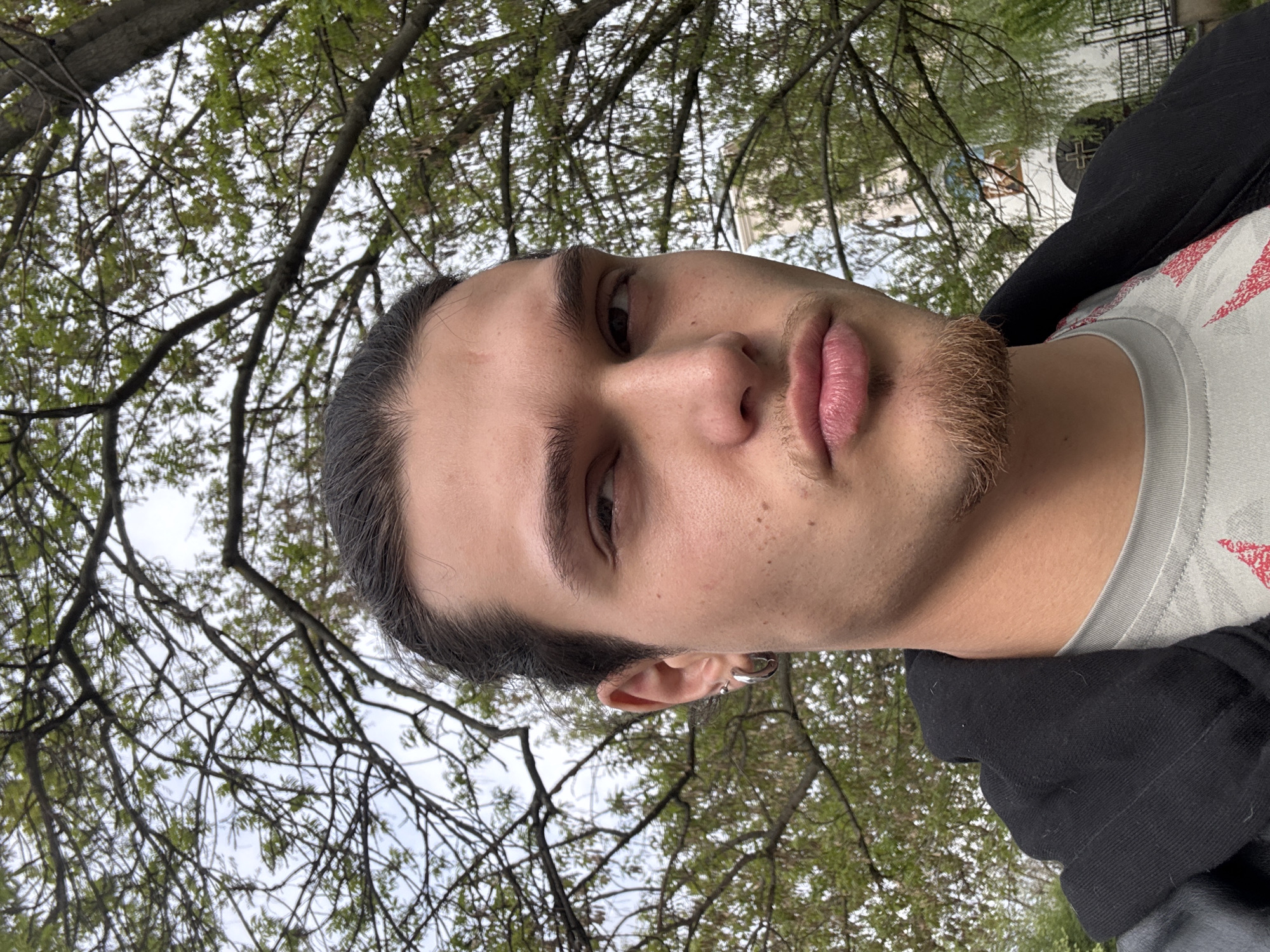 A portrait, presented vertically, captures a young person (male-presenting) from a low-angle perspective, with their face angled slightly upwards towards the camera. The subject has dark hair pulled back and features a light brown goatee and faint stubble on the upper lip. Their eyes are open and directed slightly to the viewer's left, conveying a neutral or thoughtful expression. Full, slightly parted lips are prominent. A silver hoop earring adorns the visible left ear. The person is dressed in a light-colored t-shirt featuring a red and white geometric pattern on the shoulder, worn beneath a black hooded jacket.
The background is dominated by a dense canopy of tree branches, many still bare while others show emerging green leaves, suggesting early spring. A light gray, overcast sky is visible through the foliage. In the upper right quadrant of the background, partially obscured by the trees, a white building with dark architectural details and a distinct cross-like shape can be seen, possibly indicating a church or similar religious structure in Sofia, Bulgaria. The natural light suggests an overcast daytime. No text is visible in the image.