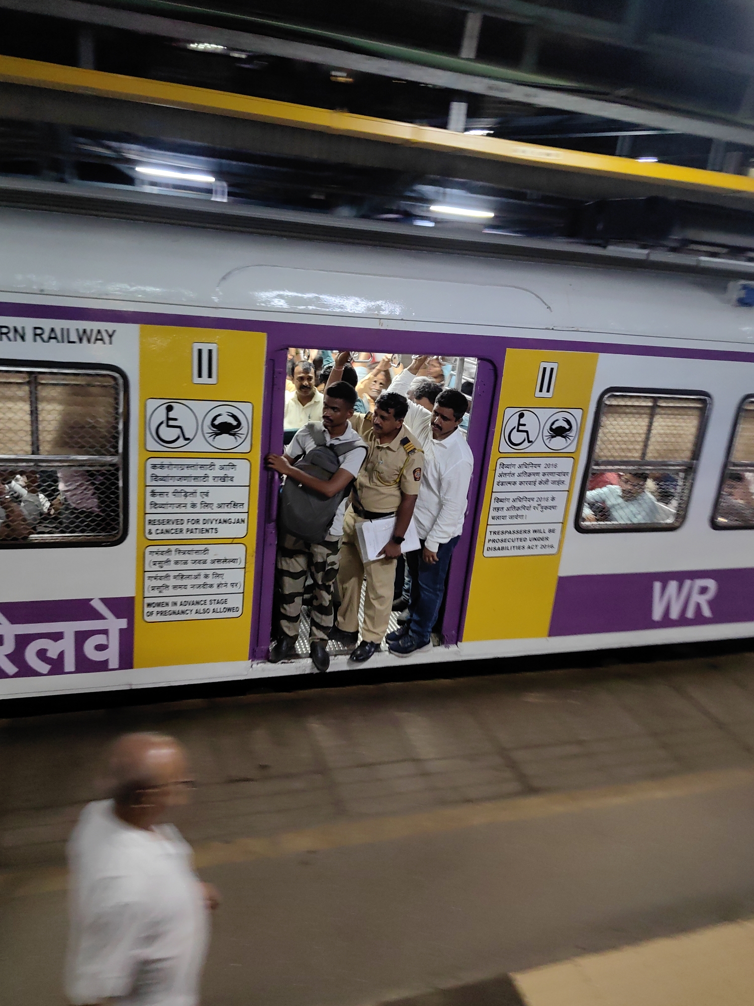 The image shows a crowded train car on the Western Railway line, likely at a station in Mira Bhayandar, India. People are packed tightly into the doorway of the train, with some standing on the platform as well. The train car is white with a purple and yellow stripe along the bottom and features signage in English and what appears to be Hindi. The signage indicates reserved seating for persons with disabilities and cancer patients, as well as for women. Two uniformed railway personnel are visible, one standing in the doorway and another on the platform. The lighting suggests it is either early morning or evening. A blurred figure of an elderly man in a white shirt is visible in the foreground, walking away from the train on the platform.