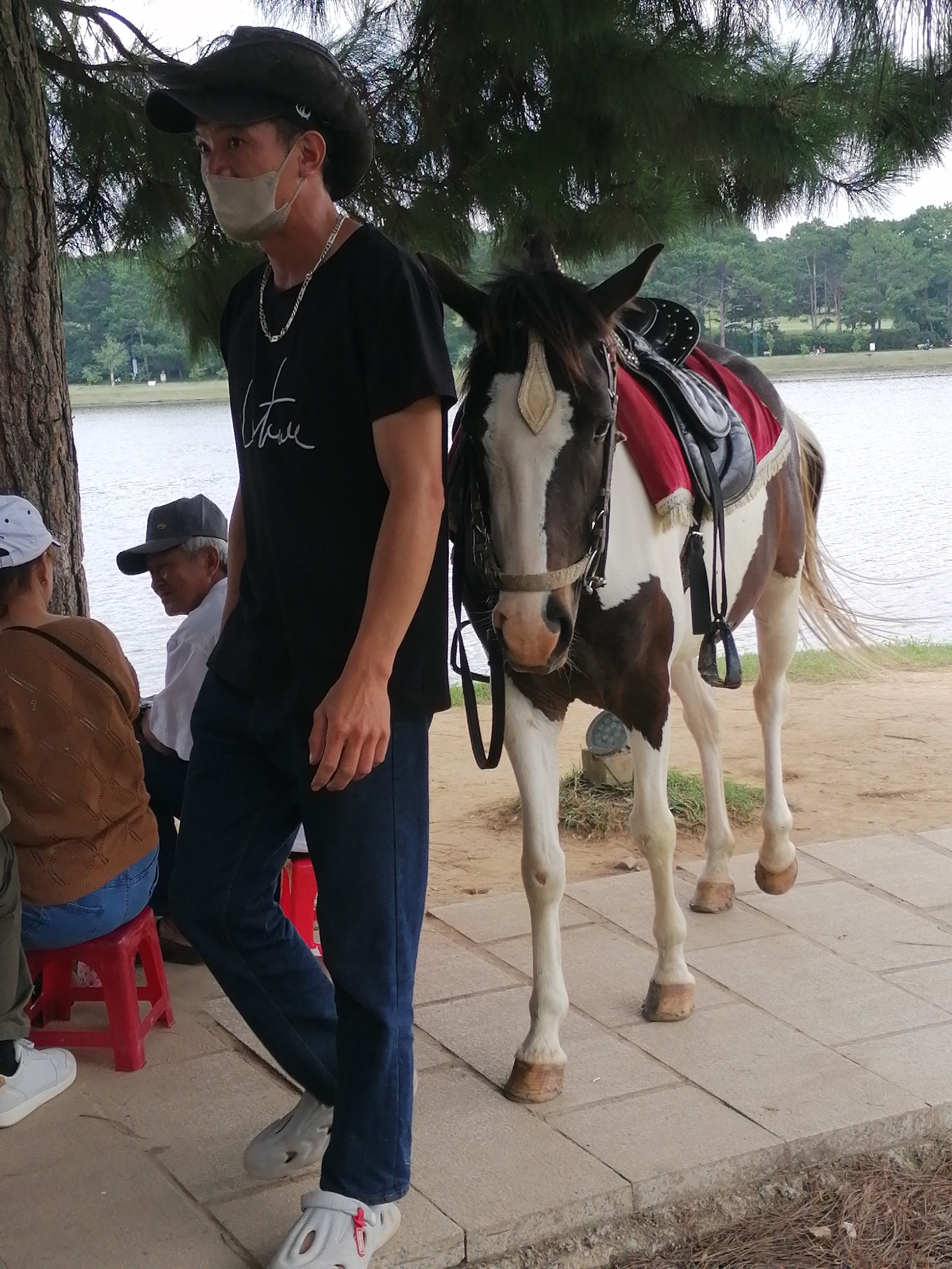 The image depicts a scene near a body of water, likely a lake, with a backdrop of trees and a slightly overcast sky, suggesting a mild day. In the foreground, a man wearing a cowboy hat and a face mask is standing next to a piebald horse. The horse is equipped with a saddle and stirrups, implying it is ready for or has just completed a ride. The man is dressed in a black t-shirt with white text, dark blue jeans, and distinctive white clogs.
To the left of the man, two other individuals are visible. One, wearing a white cap and a brown knitted sweater over a blue shirt, is seated on a red stool. Another person, a man with a grey baseball cap and a white shirt, is partially visible behind the woman on the stool, and appears to be smiling.
The setting appears to be a park or recreational area, with paved pathways and a dirt area near the water's edge. The presence of the horse suggests it might be a tourist attraction offering rides. The overall mood seems relaxed and casual. No specific time of day is definitively indicated, but the lighting suggests it is daytime. No legible text is discernible on the man's t-shirt in the provided crops.