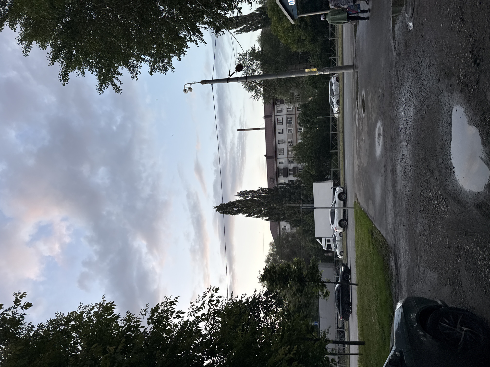 A street-level, medium-shot, eye-level photograph captures an overcast sky with scattered clouds tinged with pink and orange, suggesting either dawn or dusk. In the foreground, a dark-colored car is parked on a rough asphalt surface, with puddles reflecting the muted light. The middle ground features a wet asphalt road with several cars, including a black sedan and two white SUVs, driving or parked. A white truck is visible in the distance. Flanking the road are grassy areas and trees. Dominating the background is a multi-story apartment building with a reddish-brown roof, situated behind a line of trees. A tall, slender tree stands prominently in front of the building. A street lamp with two lights is positioned on a metal pole, with wires strung across the sky. Two figures, one wearing a green jacket and the other in a patterned dress, walk on the sidewalk. The scene appears to be a residential area in Kazan, Russia, likely during the transitional light of a day's beginning or end.