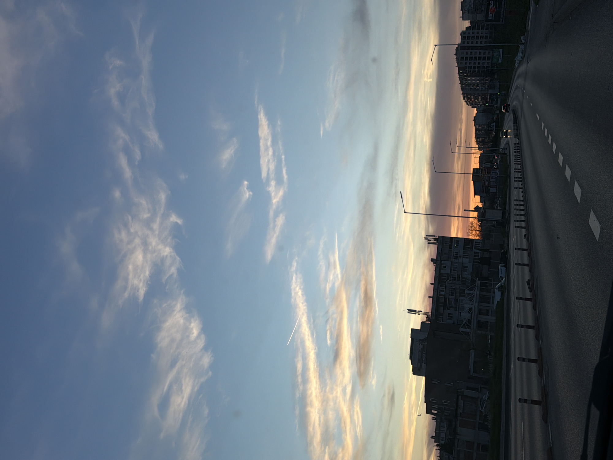 The image shows a street scene in Varna, Bulgaria at sunset. The sky is blue with streaks of white and gold clouds. A contrail is visible in the sky. On the right side of the frame, there is a dark paved road with white markings and a barrier. Buildings are present on both sides of the road, silhouetted against the sunset. Streetlights line the road. Cars are visible.