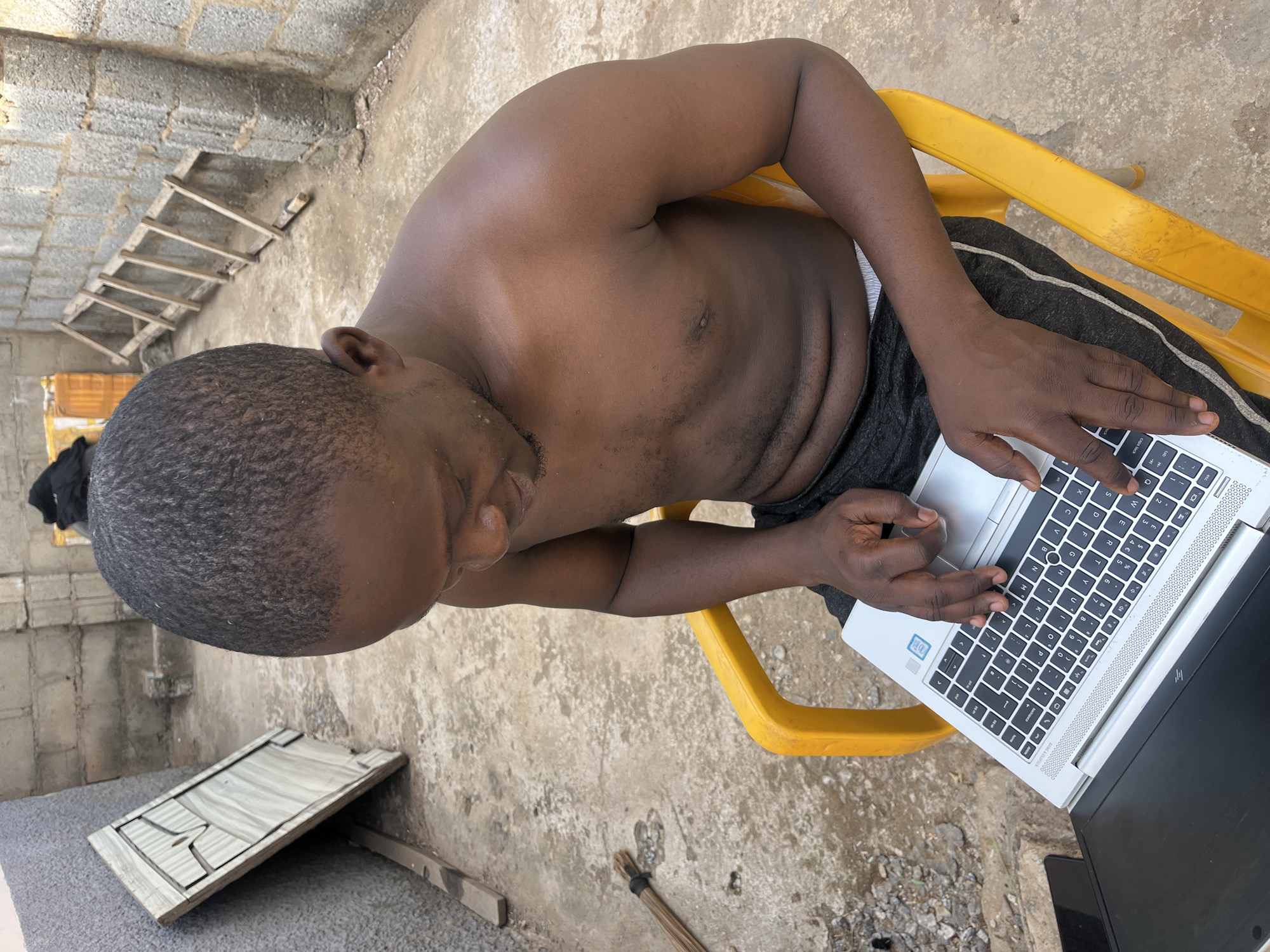 A dark-skinned man with a closely shaved head sits shirtless on a yellow plastic chair outdoors, working on a silver laptop. The location appears to be in a partially constructed area with exposed brick walls and a rough concrete floor. To the left is a wooden ladder and a damaged wooden object.