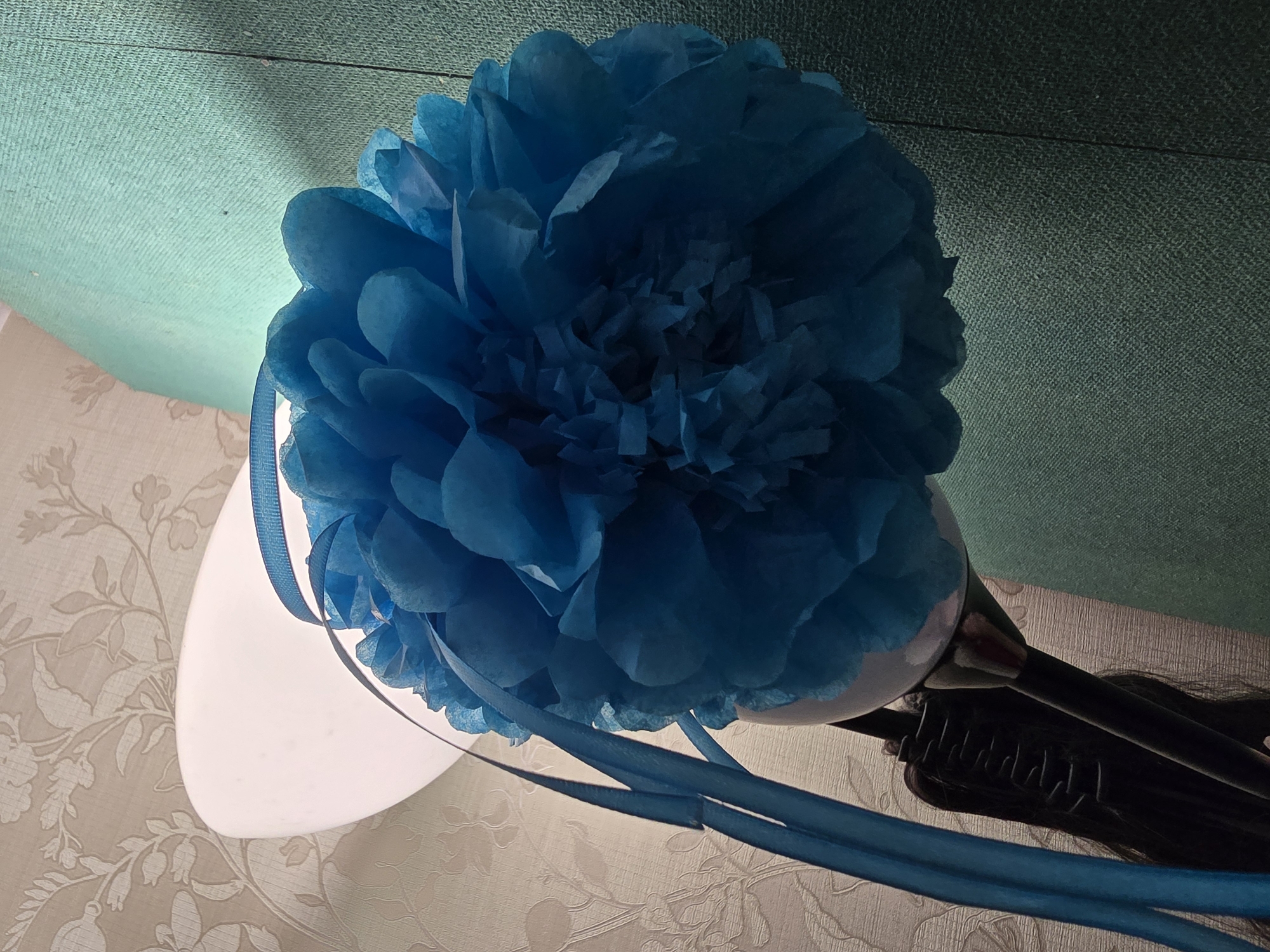 The image depicts an indoor scene, located in Krasnoyarsk, Russia, dominated by a large, spherical blue tissue paper decoration. This decoration, resembling a pom-pom or a layered paper flower, is positioned in front of a white, luminous lampshade. The lampshade emits a soft white light, backlighting the blue paper, which highlights its crinkled texture and creates a glow around its periphery. Several narrow, blue ribbons, matching the color of the decoration, are attached to it and hang downwards, some draped across the lampshade on the left.
Below the lampshade, a segment of a dark, cylindrical lamp stand is visible. Attached to this stand is a black, horizontally ribbed object, possibly a clamp or a section of the lamp's adjustable mechanism. From this area, a strand of long, dark, straight hair, consistent with a wig or hair extension, hangs downwards.
The background on the left side of the frame features light-toned wallpaper adorned with a subtle, repeating pattern of floral or botanical elements, including stylized leaves and branches. On the right, a vertical surface covered in a finely woven, textured fabric of a muted teal-green hue is visible. A clear vertical seam or panel division is present within this fabric towards the far right edge of the image. The scene is illuminated primarily by the obscured lamp, creating a contrast between the glowing decoration and the surrounding dimly lit environment.